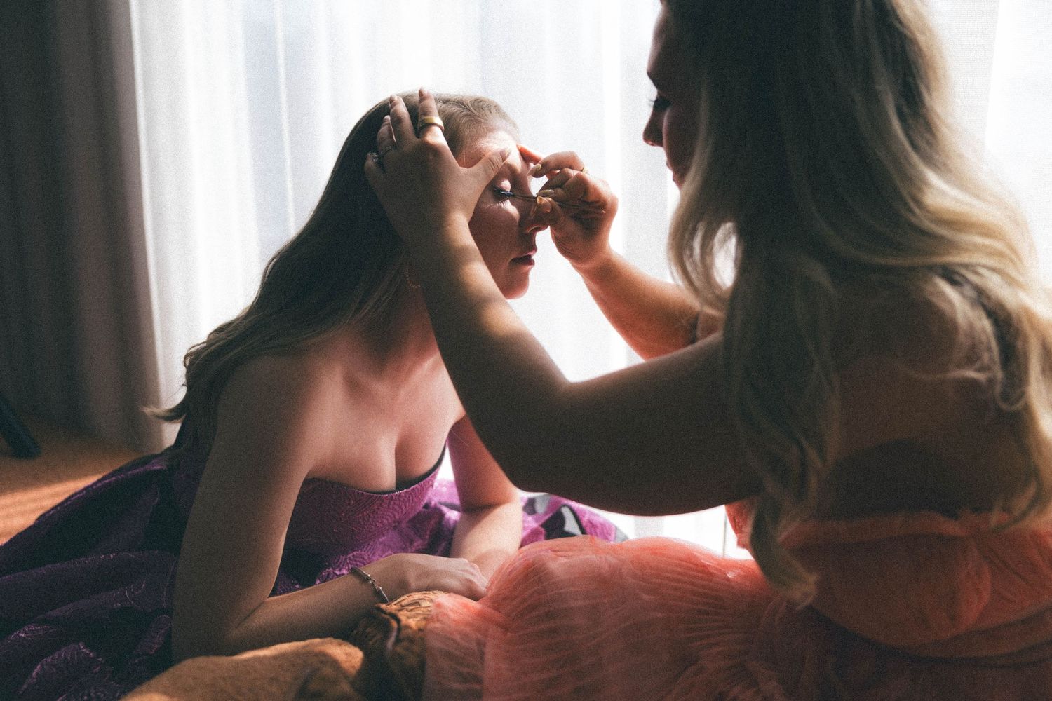 Bridesmaid helps apply makeup to bride sitting near a sunlit window during wedding preparations.