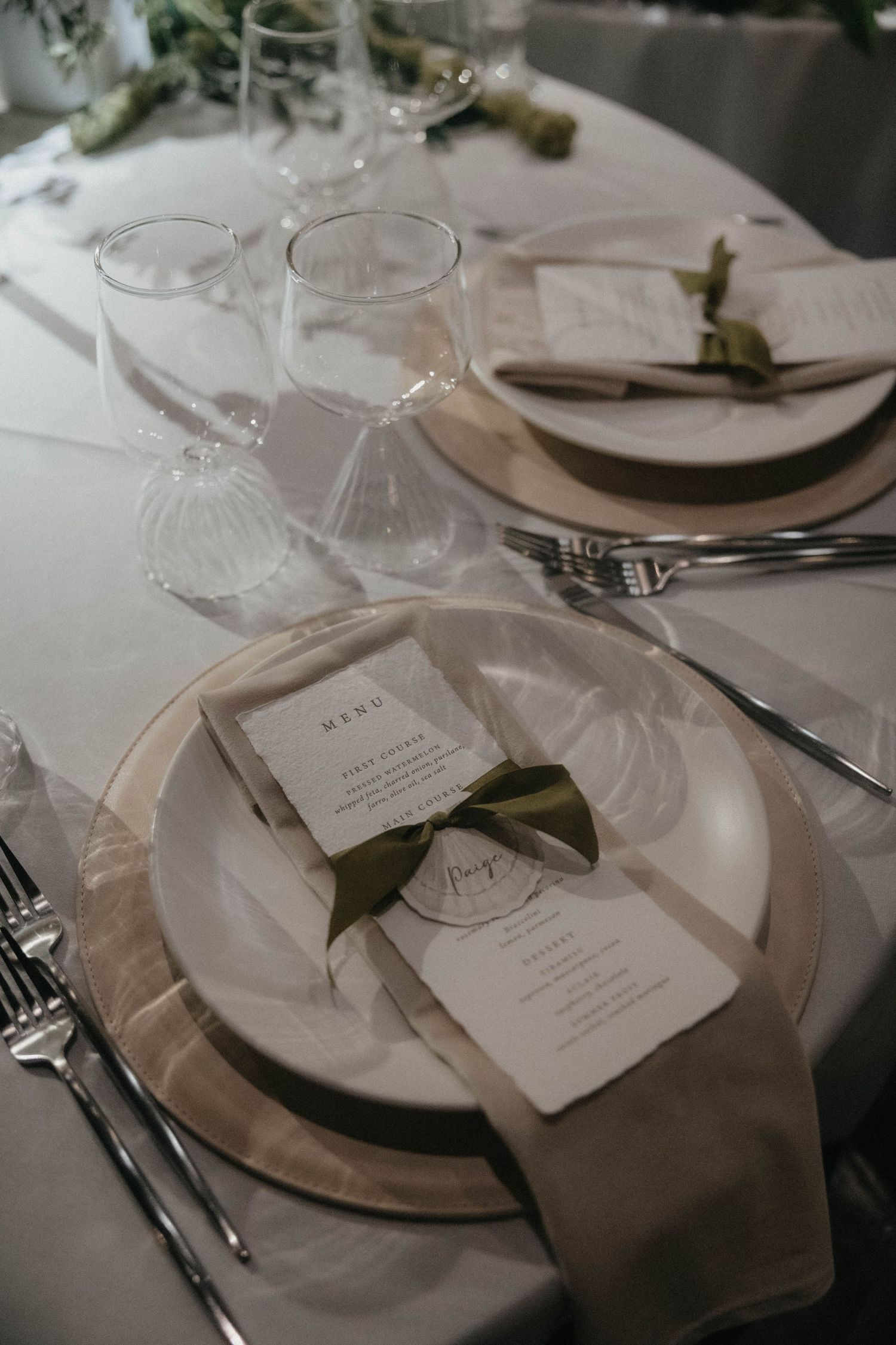 Elegant wedding table setting with a taupe linen napkin, menu card tied with an olive green ribbon, gold charger plate, and clear stemware.