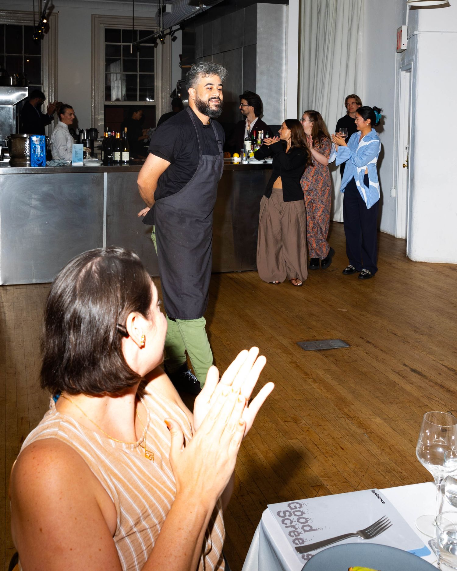 A bearded chef in a black apron stands on a wooden floor as guests applaud and a woman in the foreground claps at a lively dinner event.