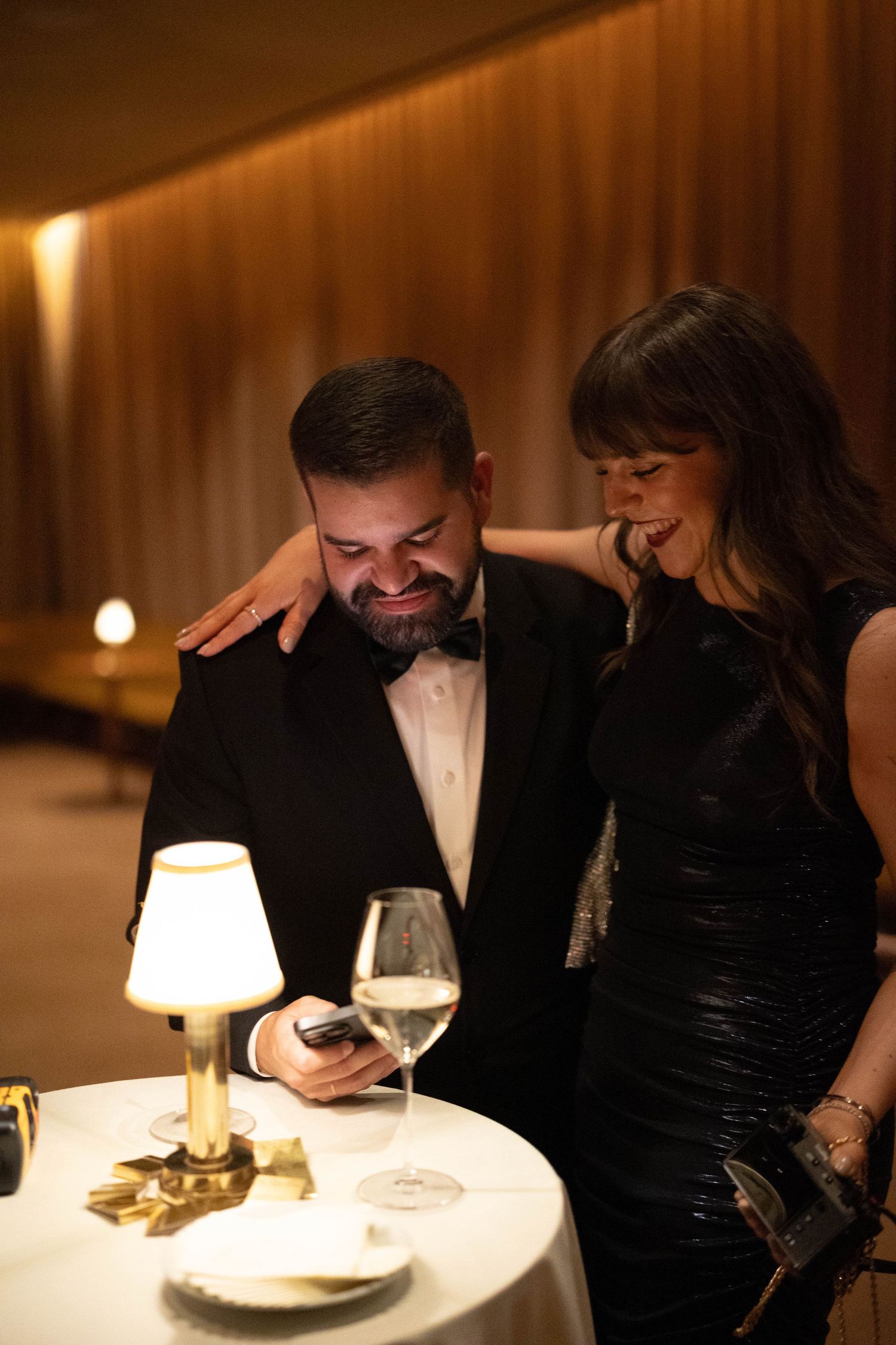A smiling couple in formal attire enjoys an elegant evening at a candlelit restaurant table with a lamp and a glass of white wine.
