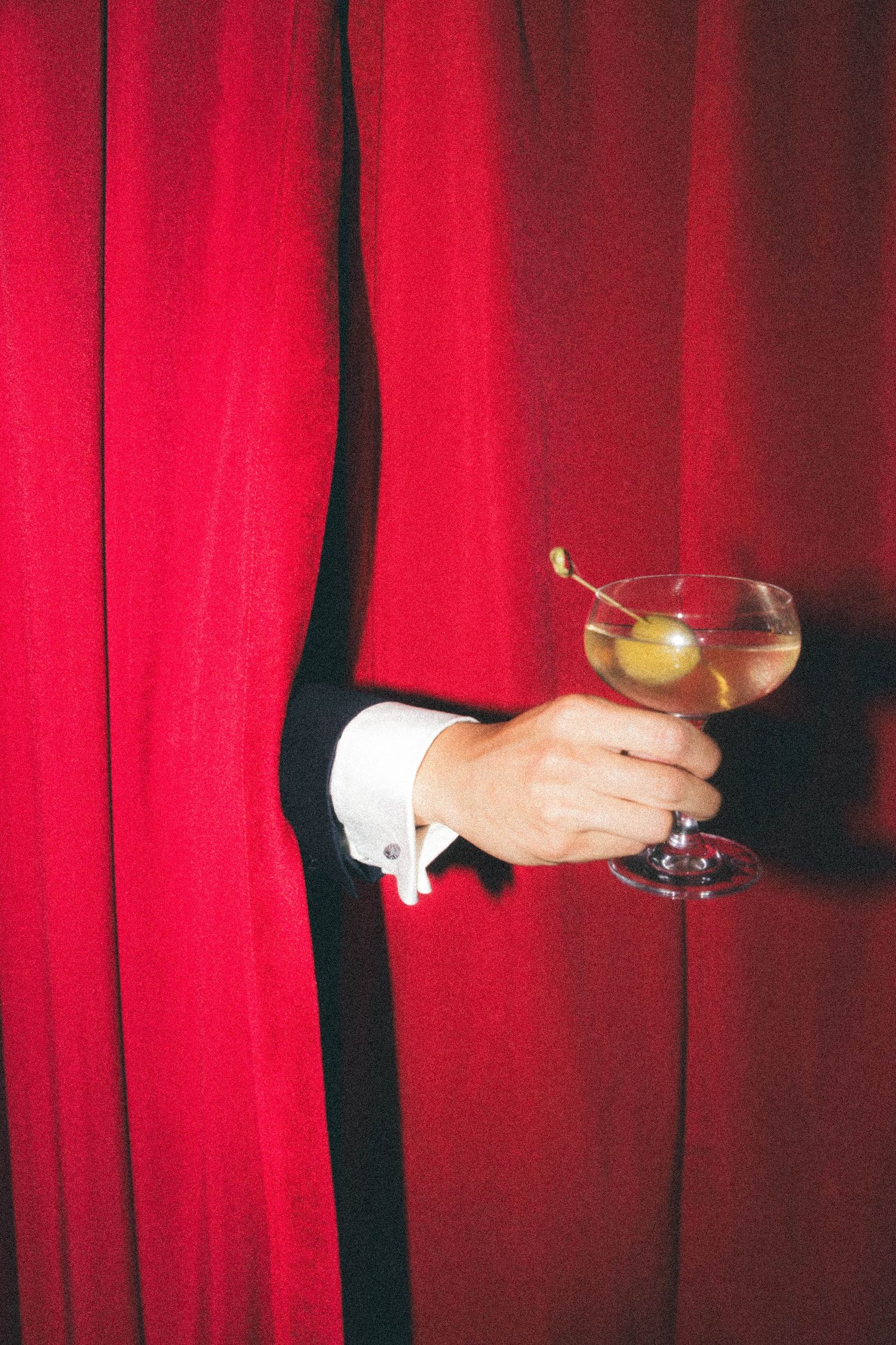 A hand emerging from red velvet curtains holding a martini glass with olive garnish.