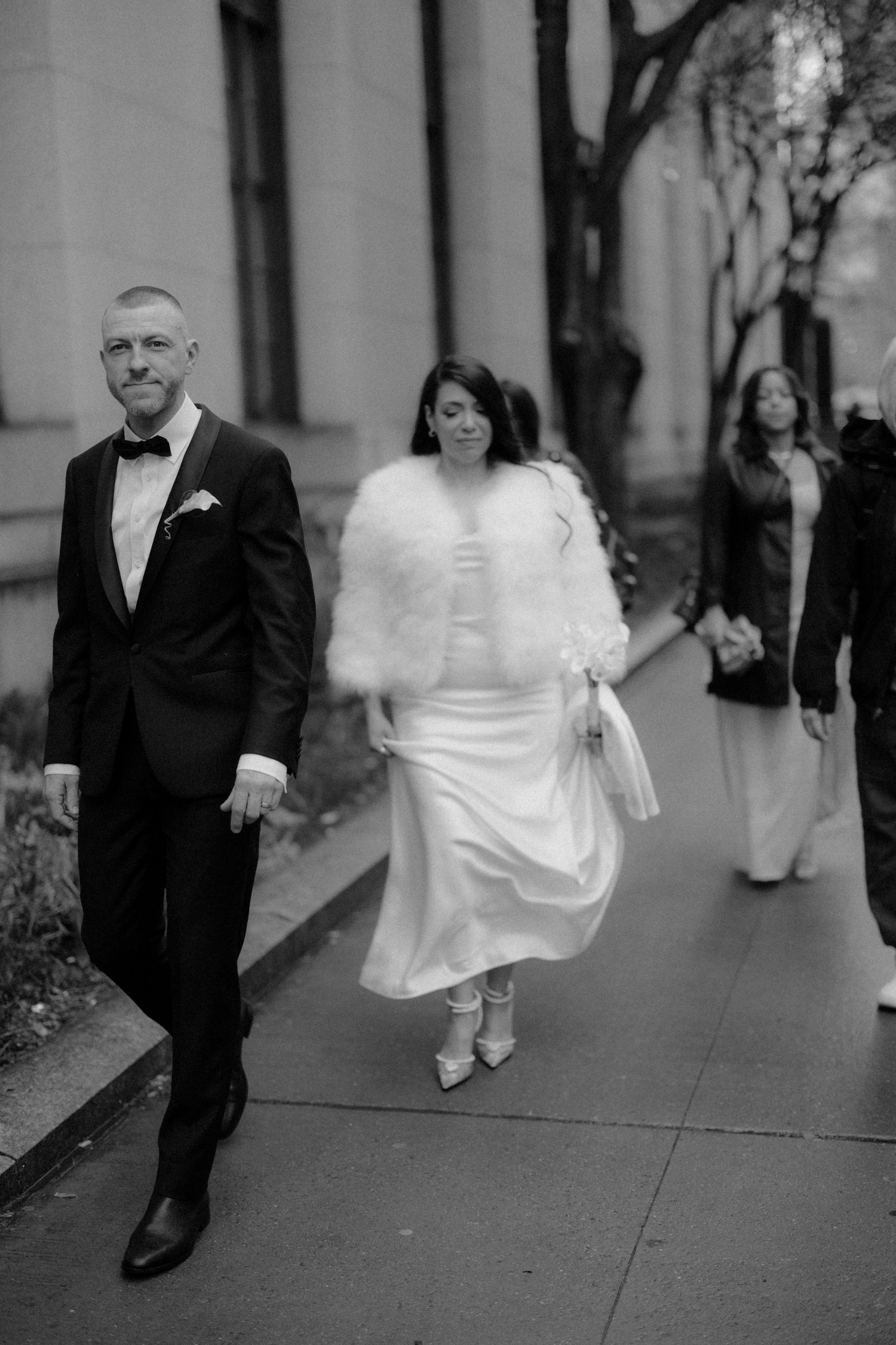 Bride in white gown with fur stole and groom in tuxedo walk outdoors after wedding.