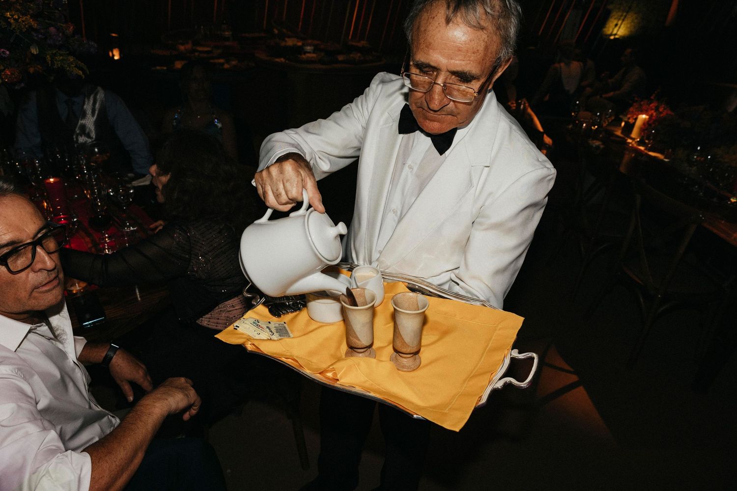 Elderly waiter in white jacket pouring drinks from pitcher onto tray at wedding reception.