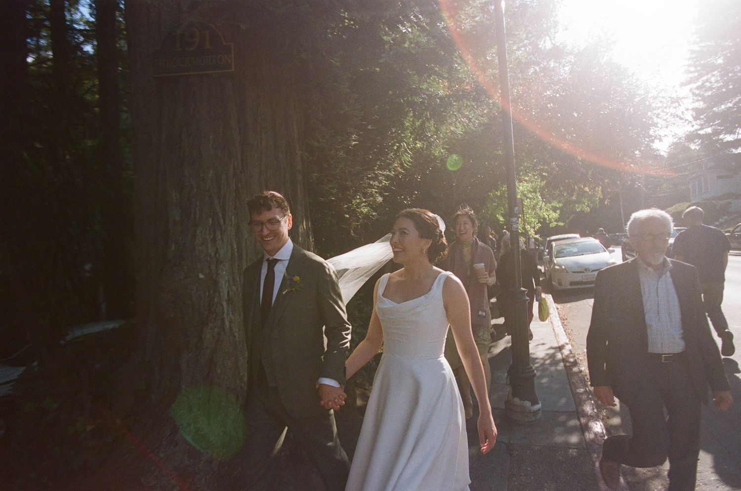 Bride and groom walk in golden sunlight along a tree-lined street with guests.