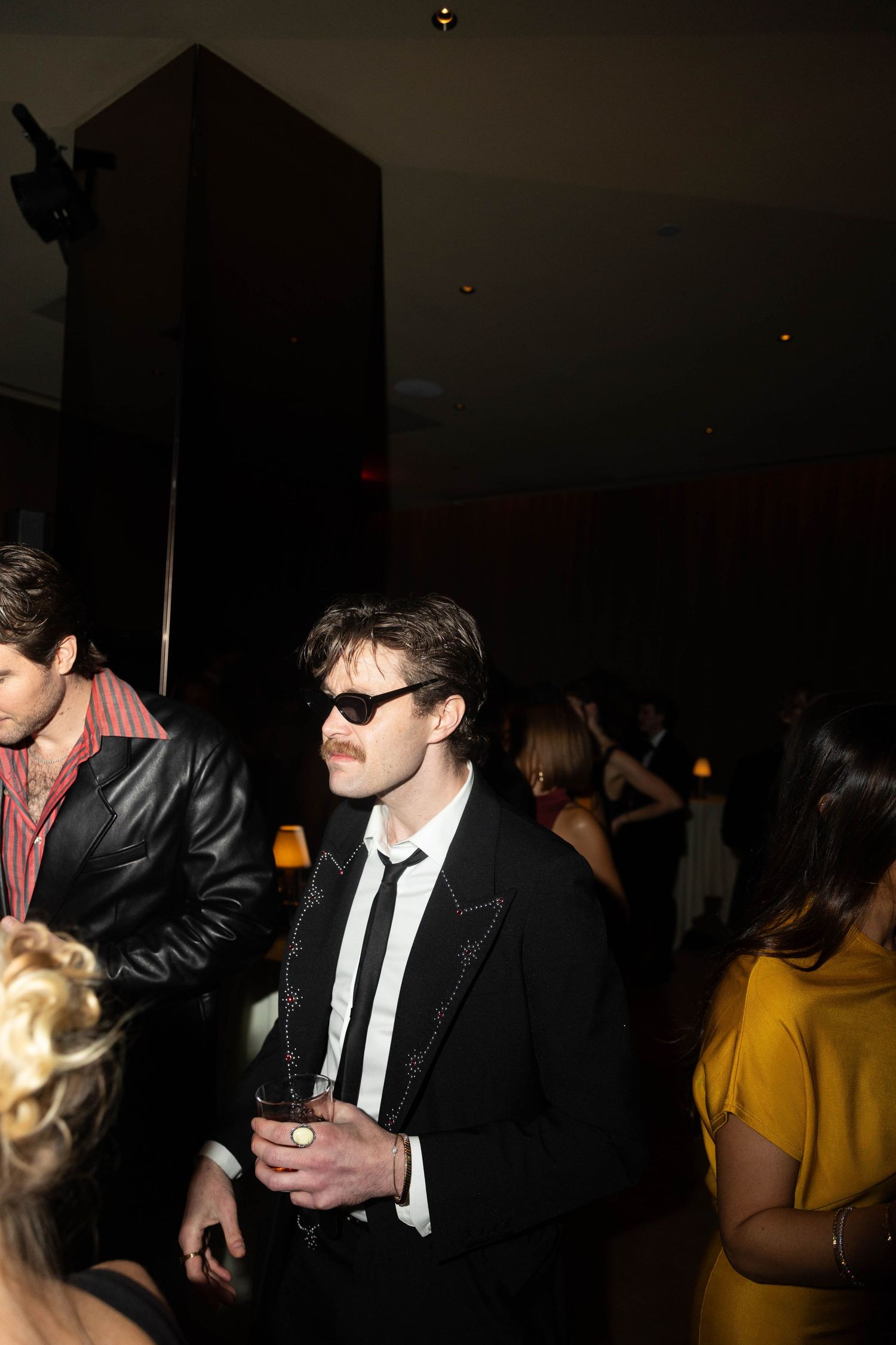 Man with a mustache and sunglasses wears a black suit with satin trim and holds a drink at a dimly lit party with guests behind him.