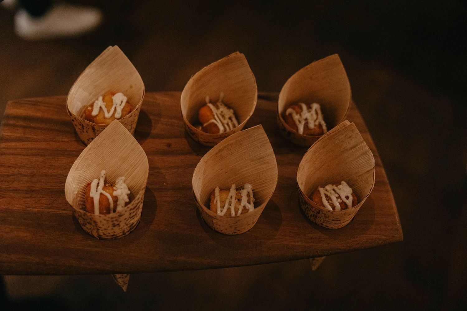 Six small wooden cones filled with appetizers drizzled with white sauce, served on a rustic wooden serving board.