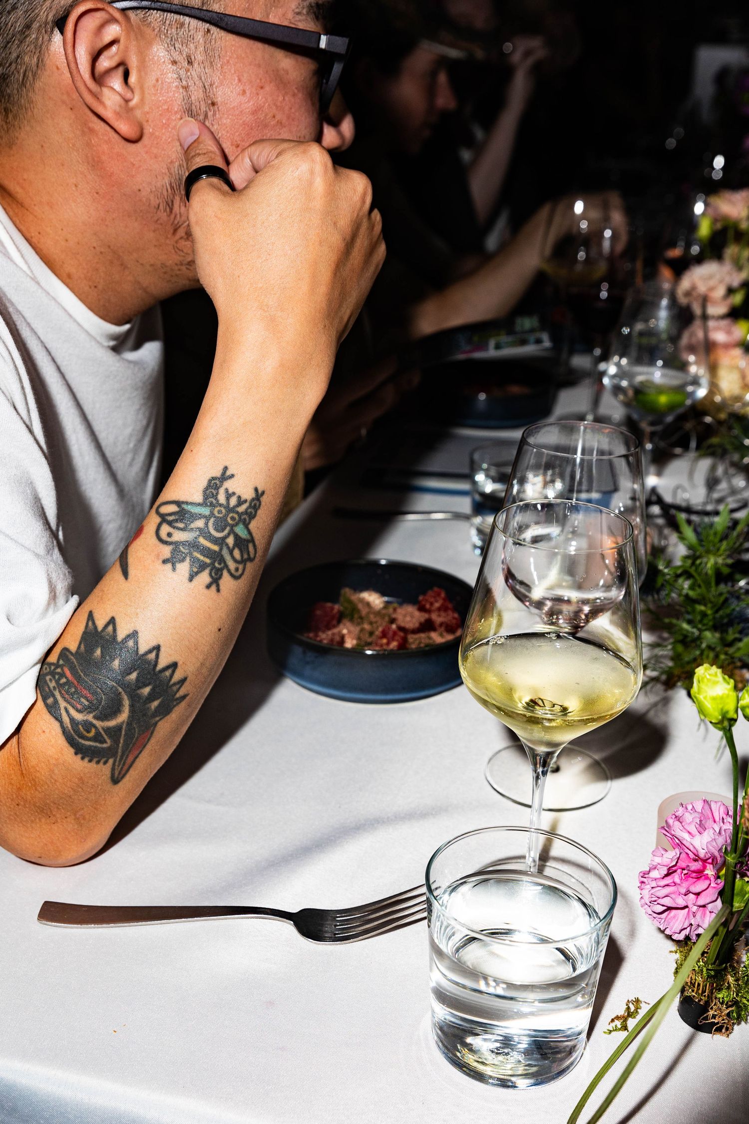 A tattooed man rests his chin on his hand at a dinner table set with wine glasses, water, a plated dish, and fresh flowers.