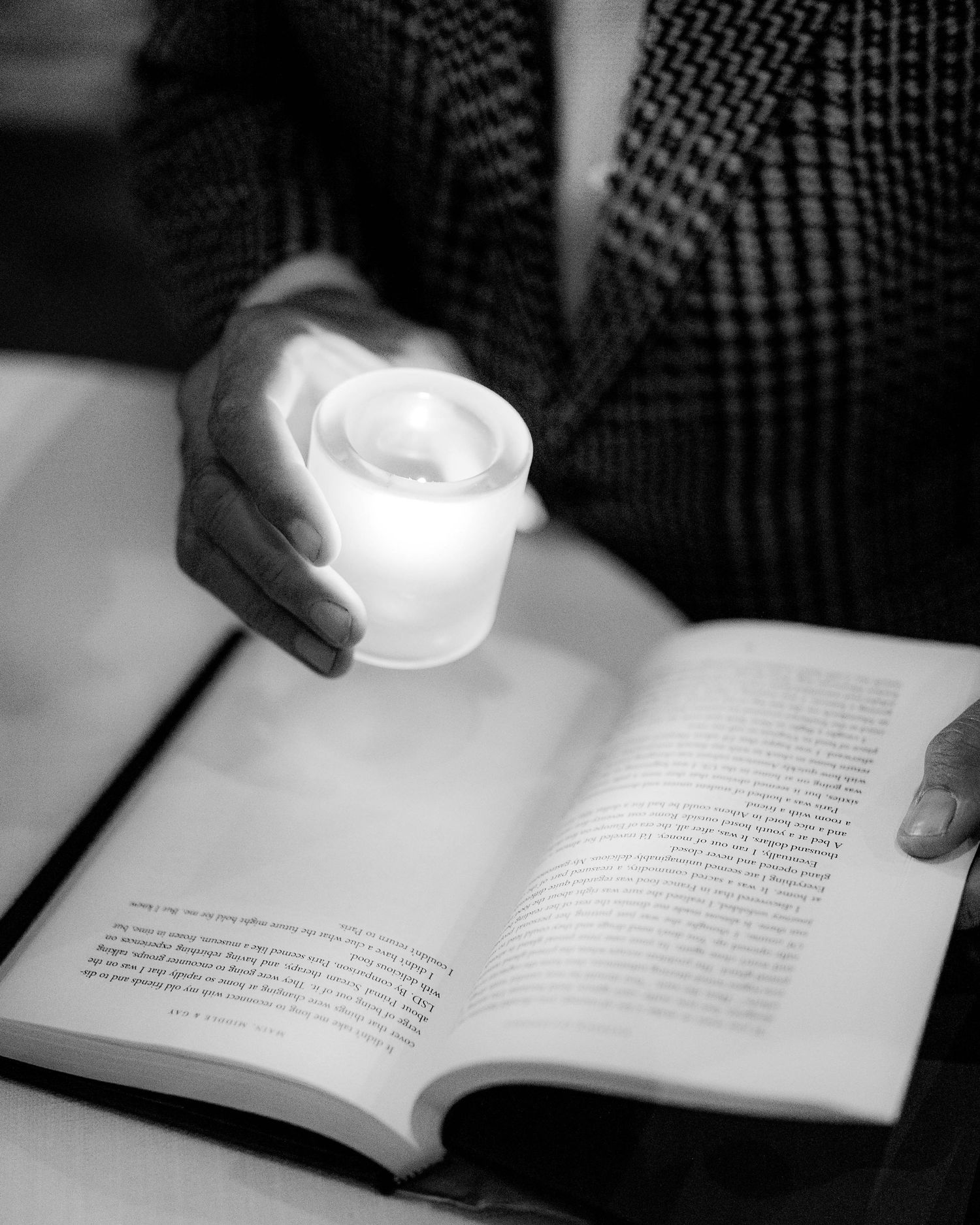 Person in a patterned blazer holds a glowing candle over an open book, reading by soft candlelight in a black and white photo.