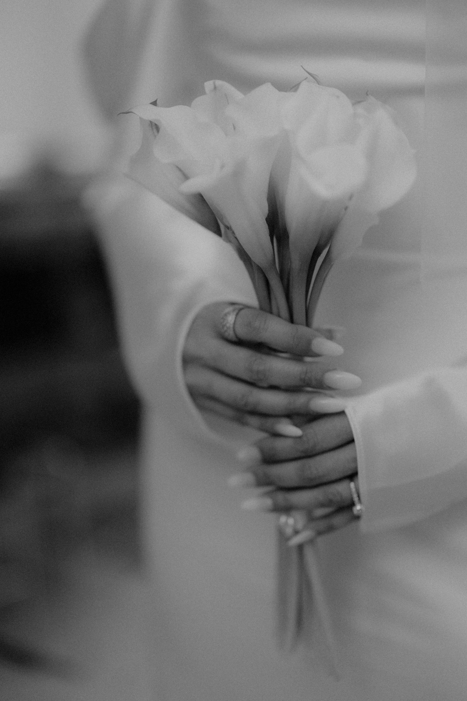 Black and white close-up of bride holding elegant calla lily bouquet with diamond ring visible.