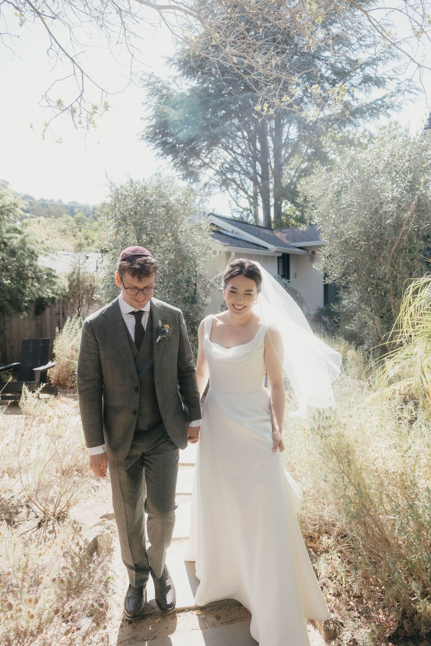 Bride and groom walk hand in hand along a sunny garden path, veil flowing in the breeze.
