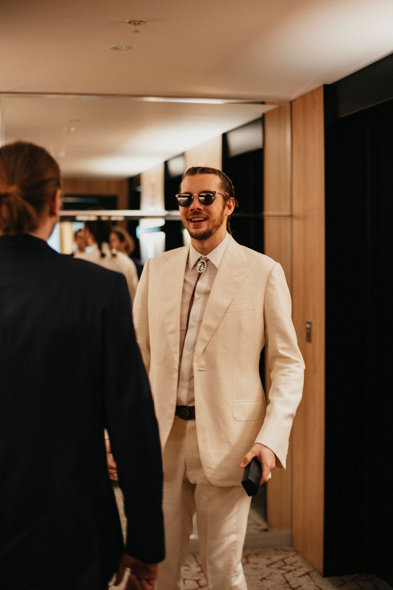 Stylish groom in cream suit and sunglasses smiling in hotel corridor before wedding ceremony.