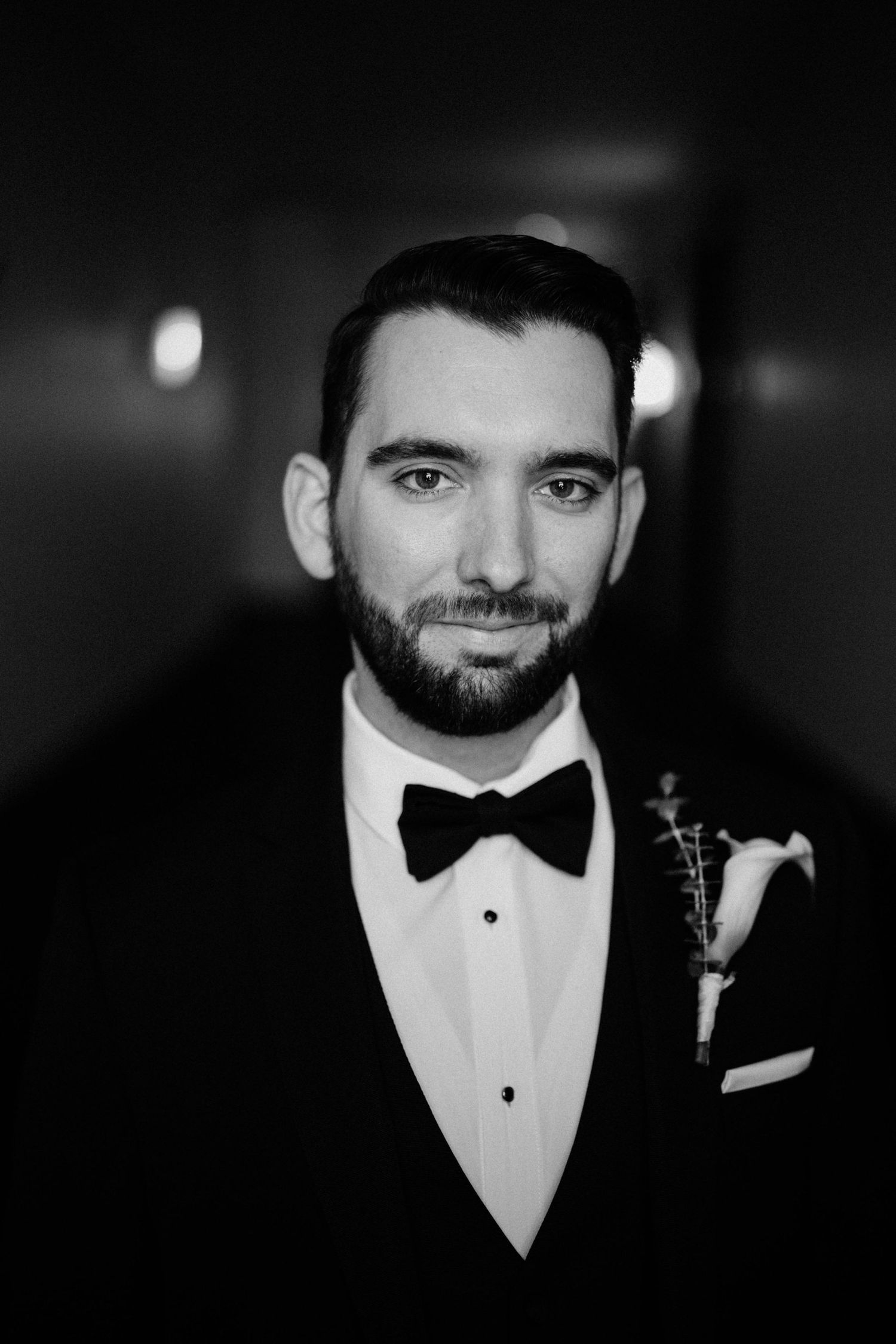 Black and white portrait of bearded groom in tuxedo with bow tie and boutonniere in a hallway.