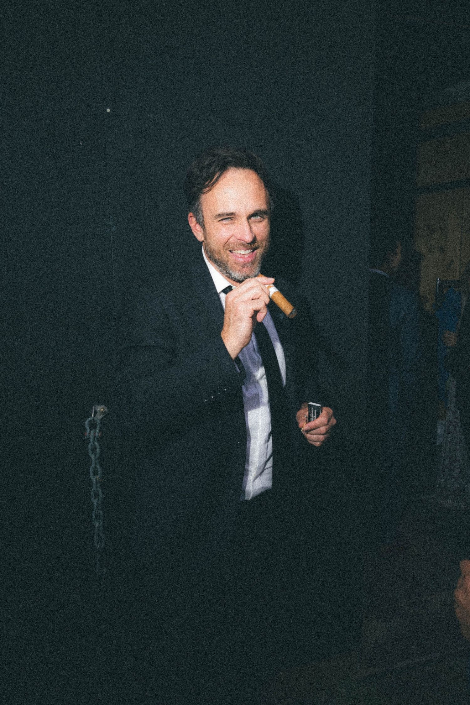 A smiling man in a dark suit holds a cigar at an evening wedding reception.