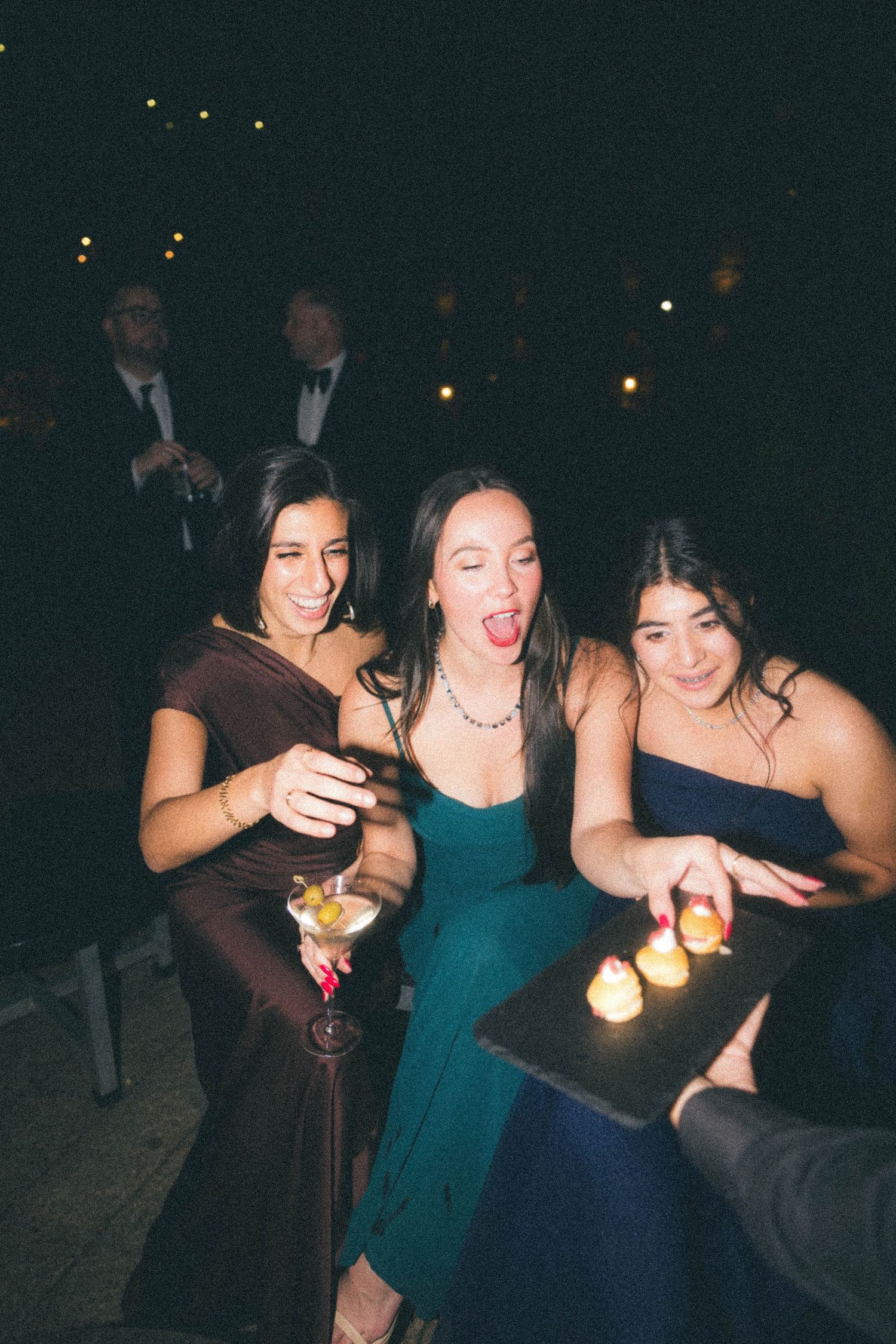 Three women in elegant gowns excitedly reach for lit canape appetizers at a formal event.
