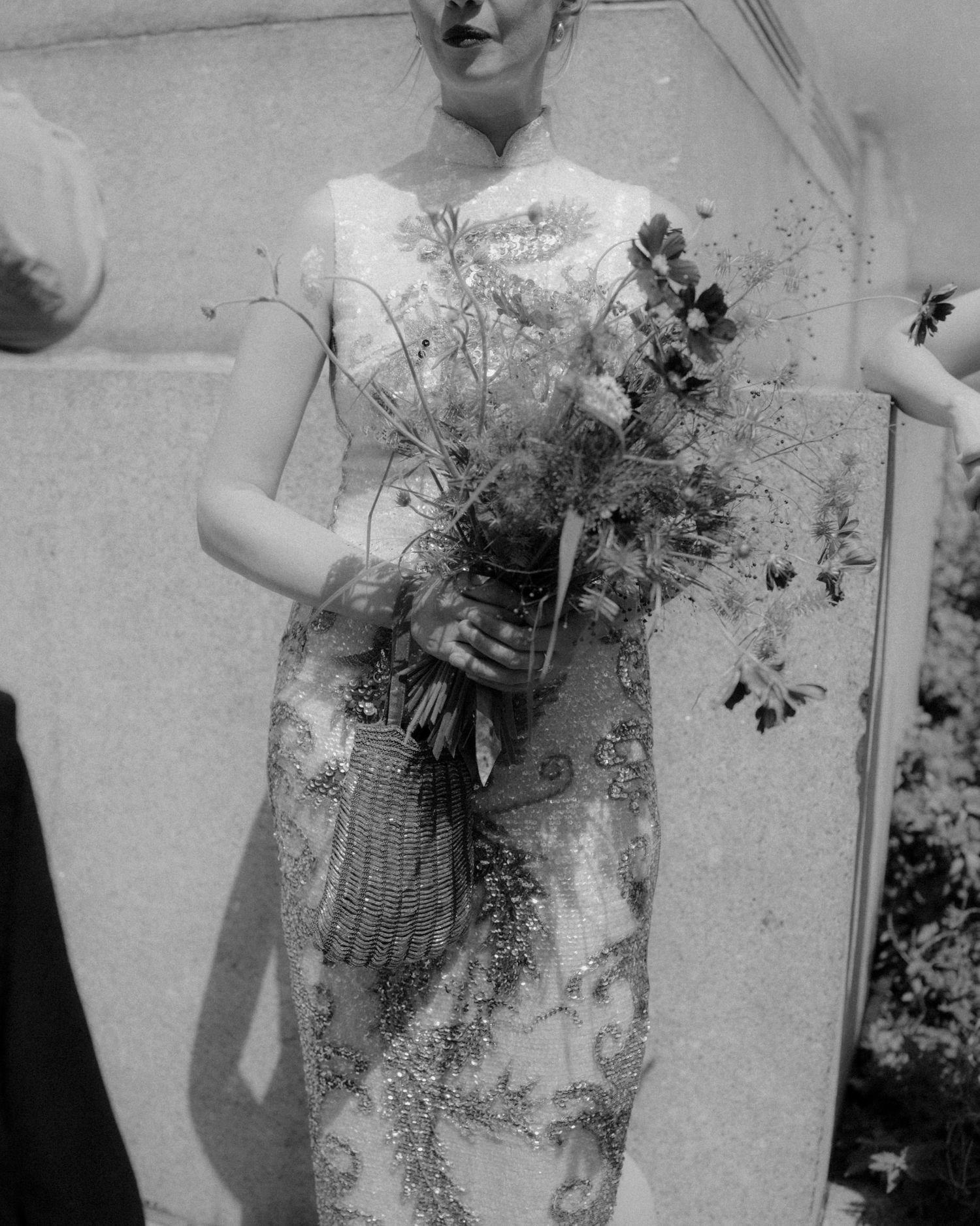 Black and white close-up of bride holding wildflower bouquet in ornate beaded wedding gown outdoors.