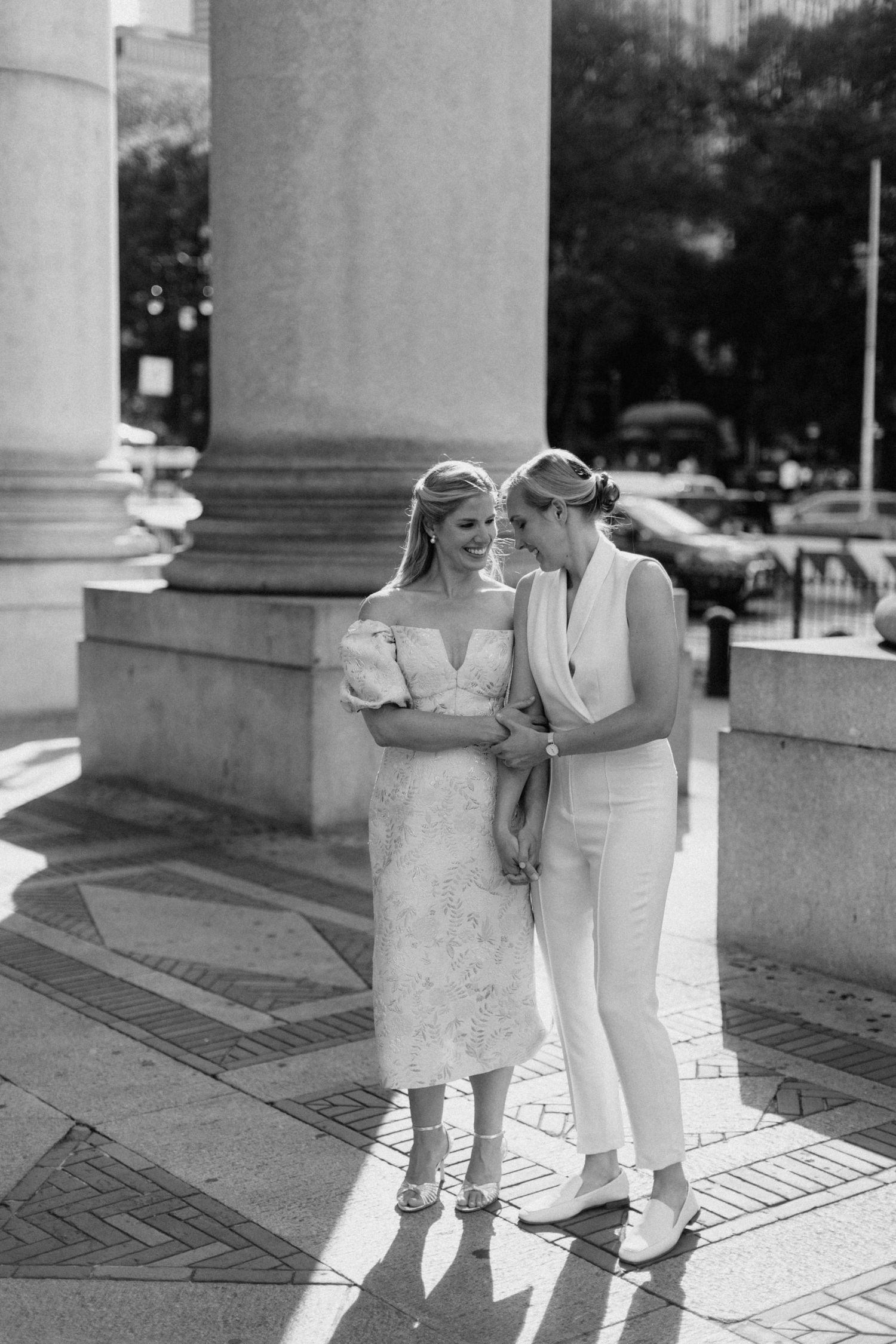 Black and white photo of two brides laughing together beside grand stone columns outdoors.