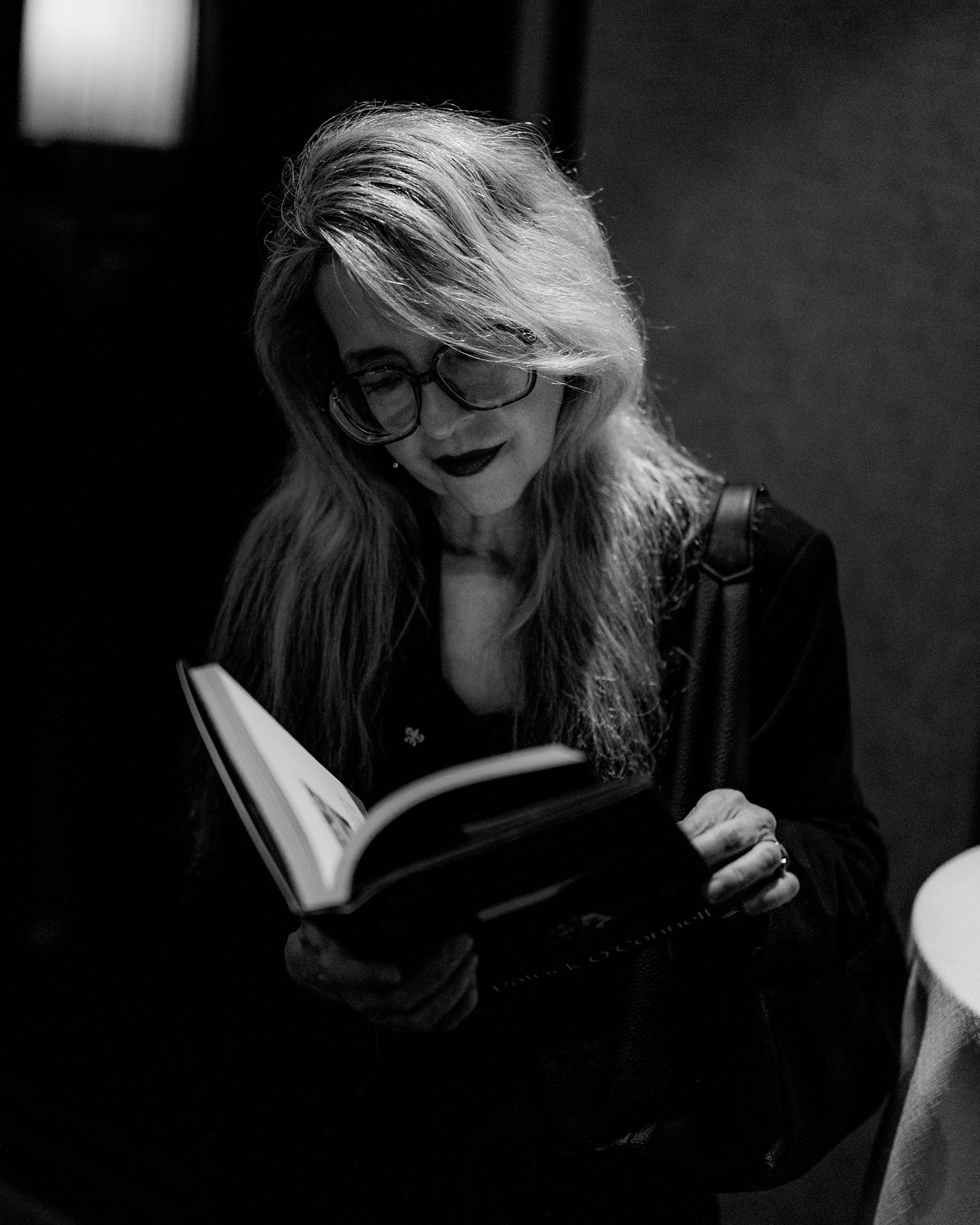 Black and white portrait of a woman with long hair and glasses reading an open book while carrying a shoulder bag.