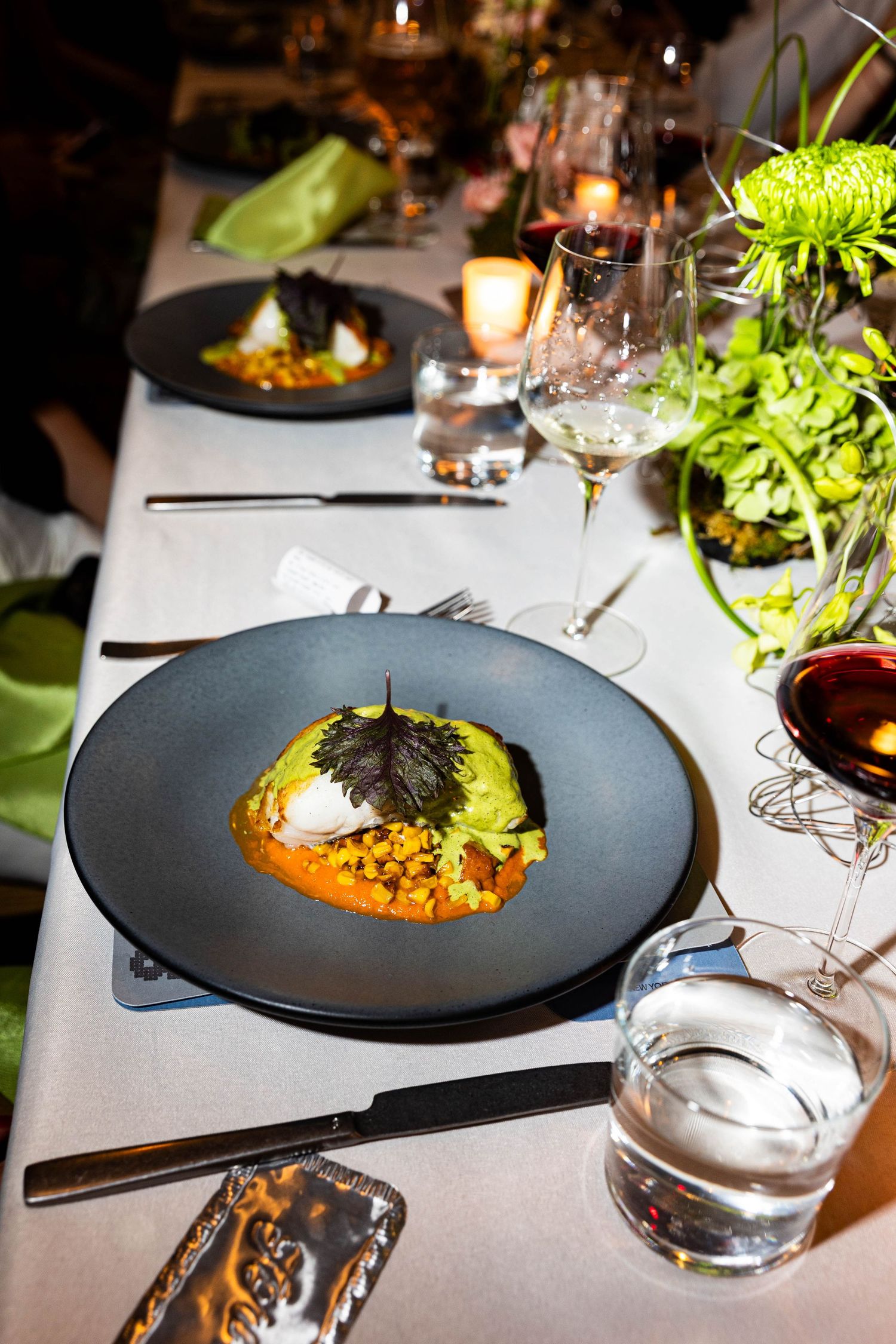 Gourmet entree with herb garnish on a dark plate at an elegant candlelit dinner table with wine glasses and fresh green floral centerpiece.