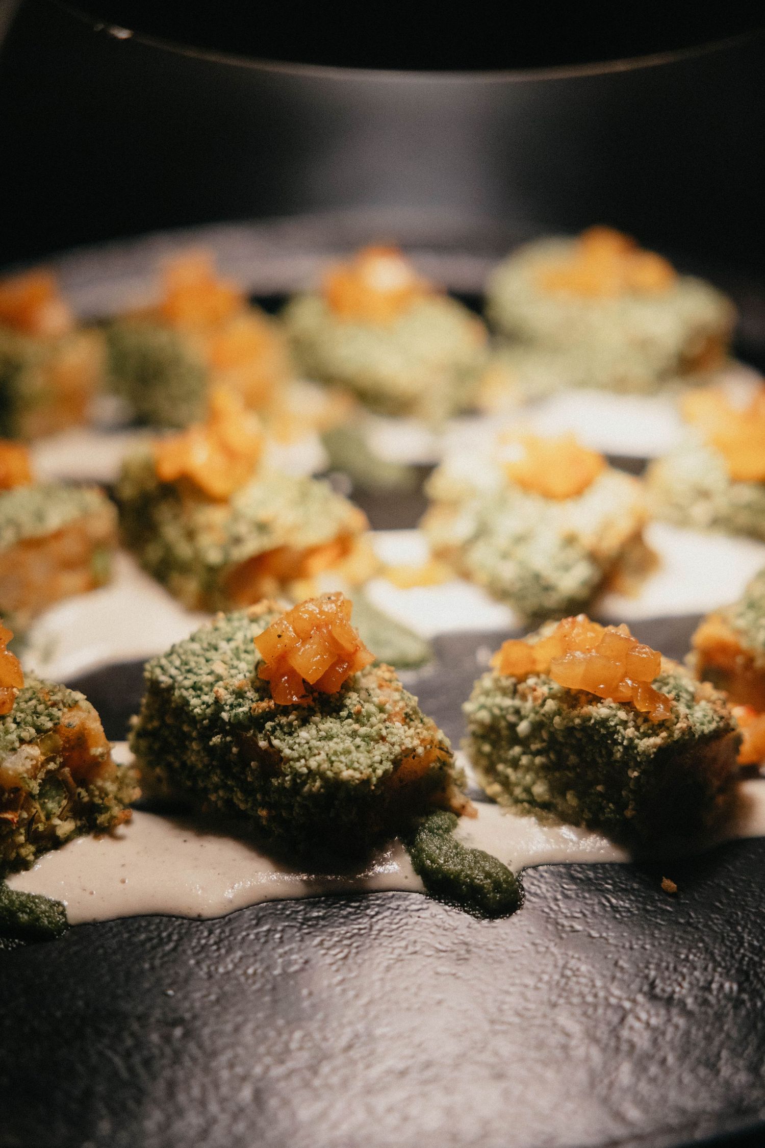 Herb-crusted green bites topped with diced orange garnish and served on creamy white sauce atop a dark plate.