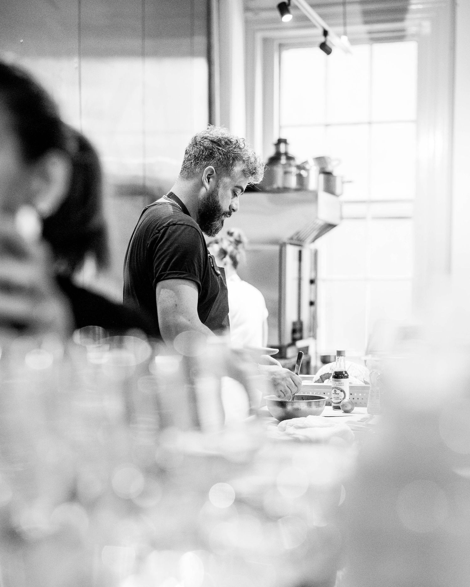 A bearded chef in a dark t-shirt works at a busy kitchen counter in a bright restaurant, shown in black and white.