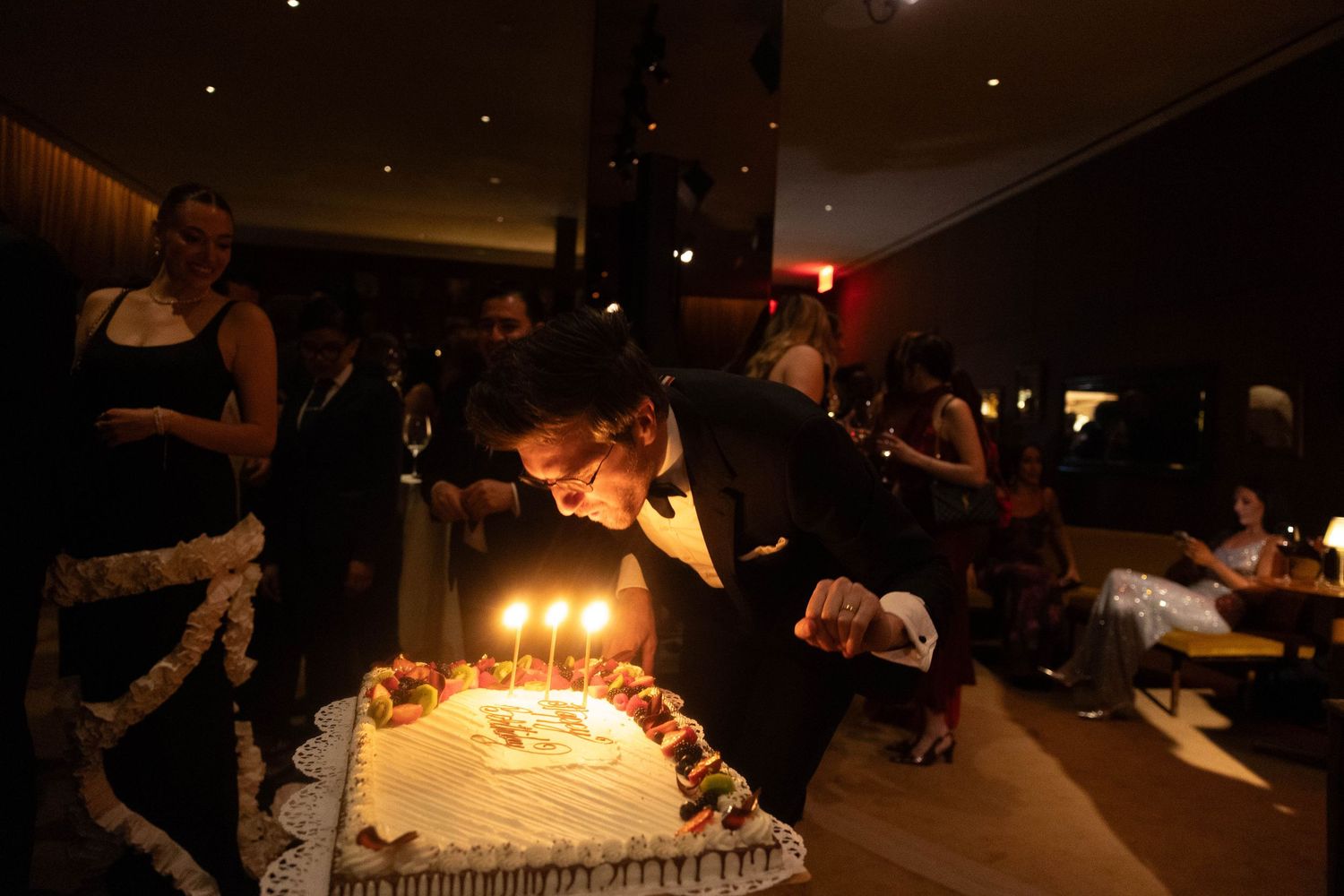 Man in a dark suit leans over a decorated birthday cake with lit candles while guests look on at an evening party in a dimly lit room.