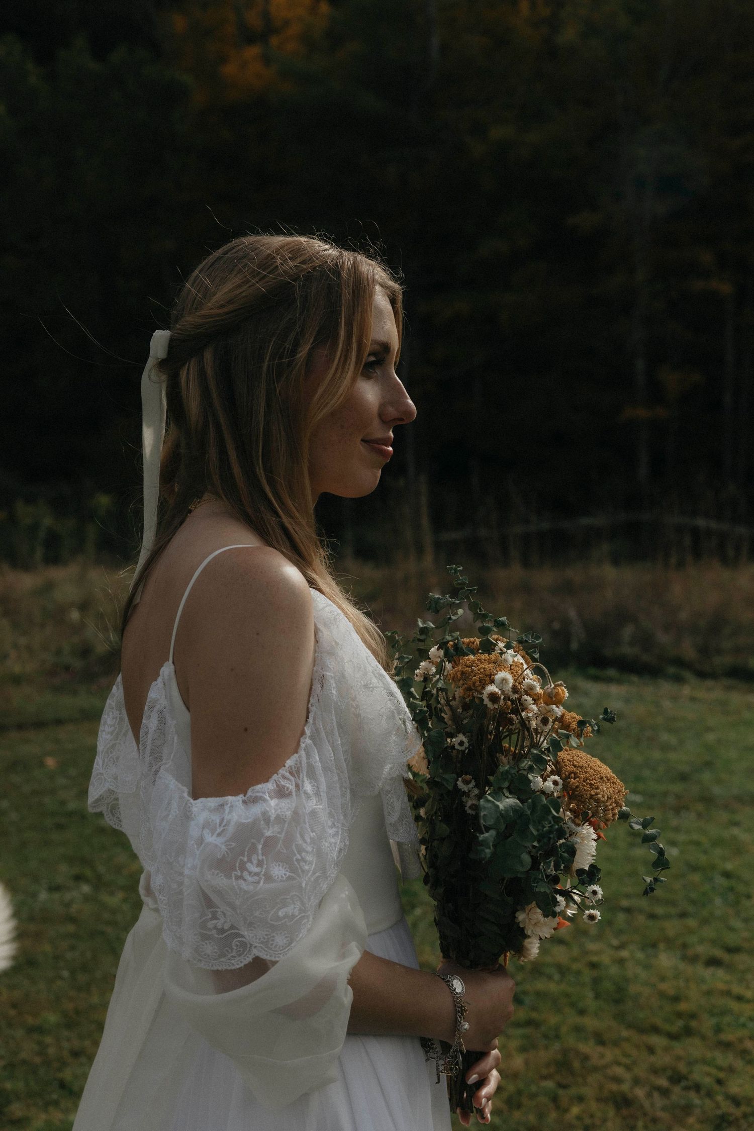 Bride in white lace off-shoulder gown holding wildflower bouquet in autumn forest setting.