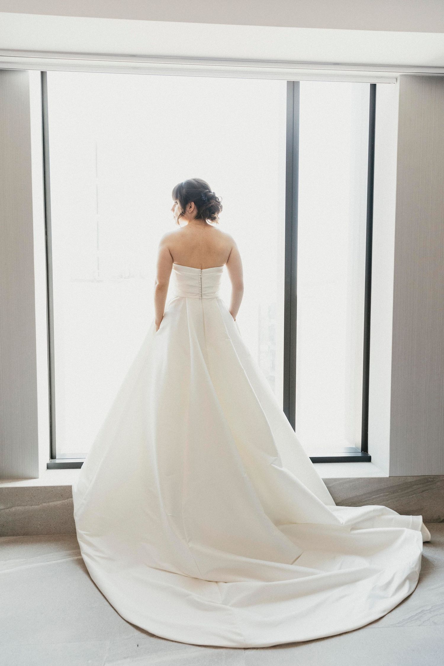 Elegant bride in a strapless white ballgown with long train stands before bright windows on wedding day.
