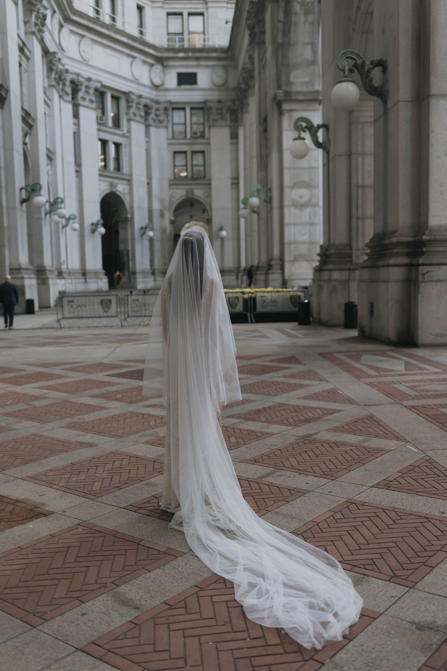 Bride in flowing cathedral veil stands alone in ornate courtyard of grand classical stone building.