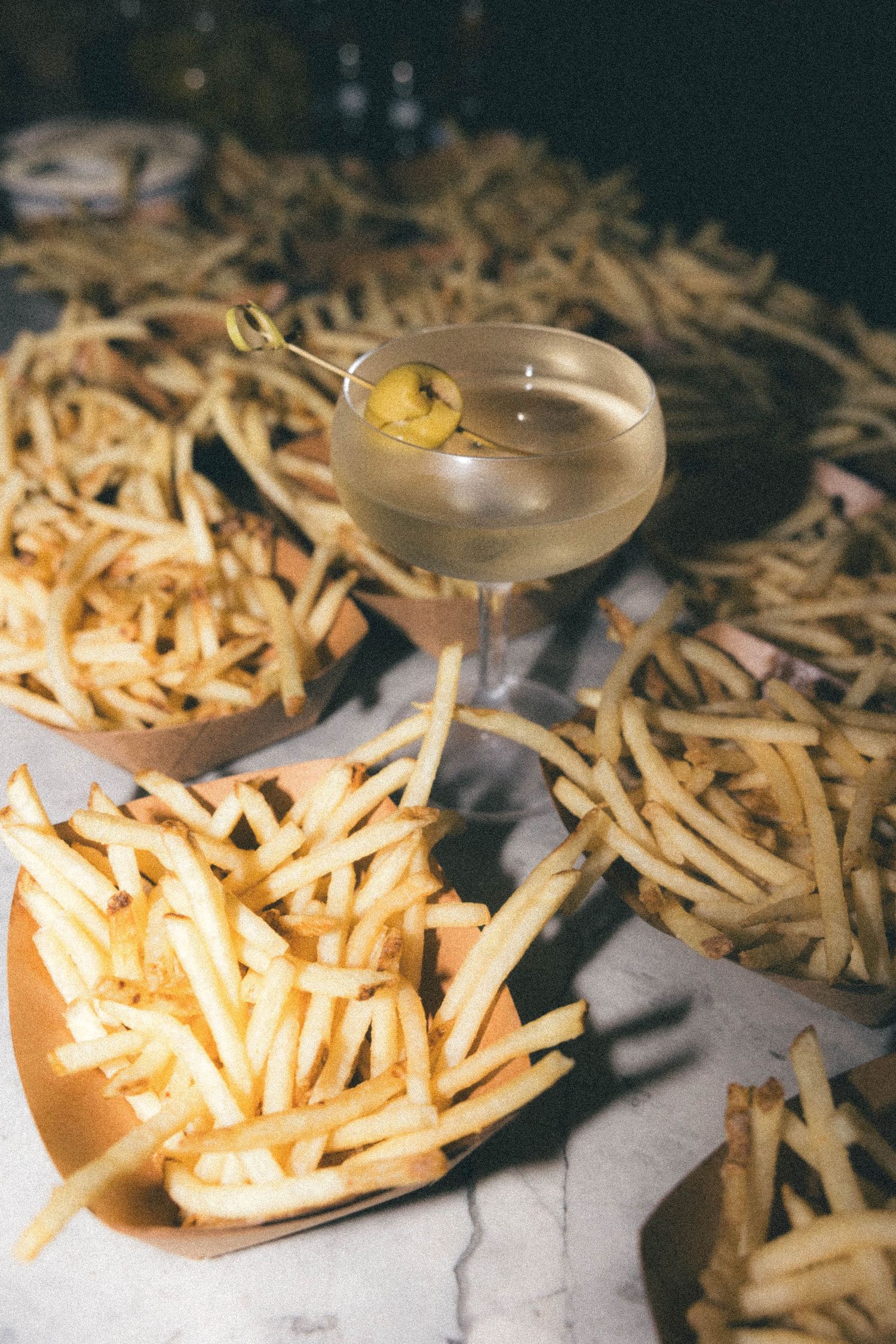 Martini glass with olive garnish surrounded by baskets of french fries on marble surface.