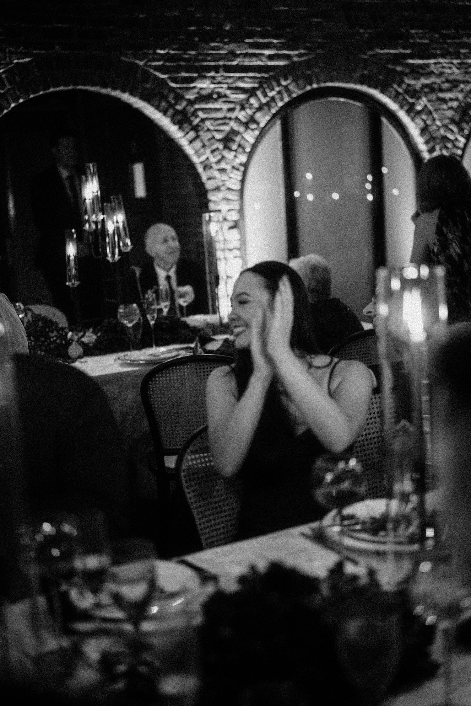 Wedding guest in black dress laughing with hands on face at candlelit dinner reception.