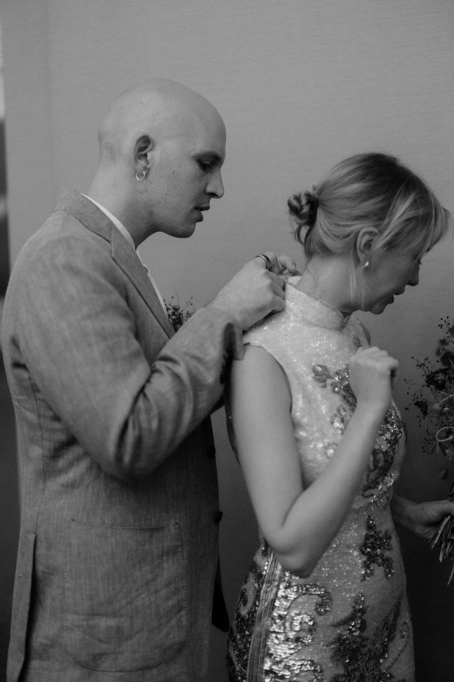 Black and white photo of groom carefully fastening bride's sequined dress before ceremony.