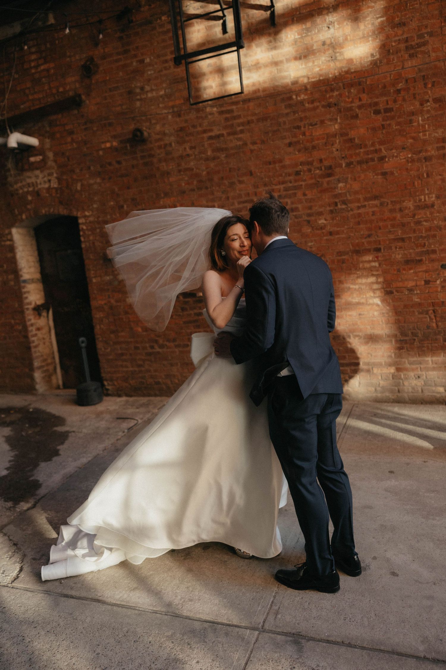 Bride and groom embracing tenderly against rustic brick wall with veil flowing in the breeze.