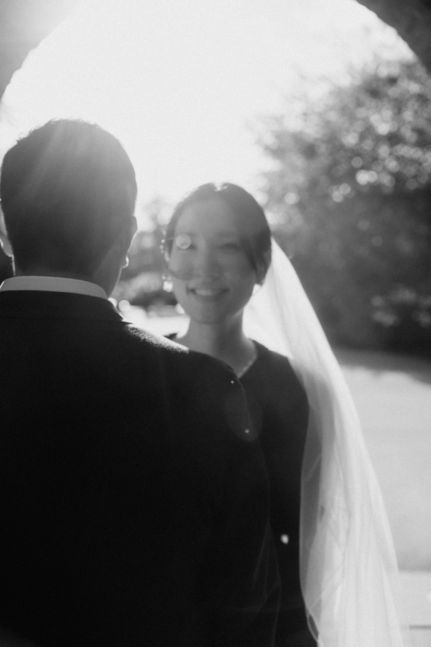 Black and white backlit portrait of smiling bride in veil facing groom at wedding.