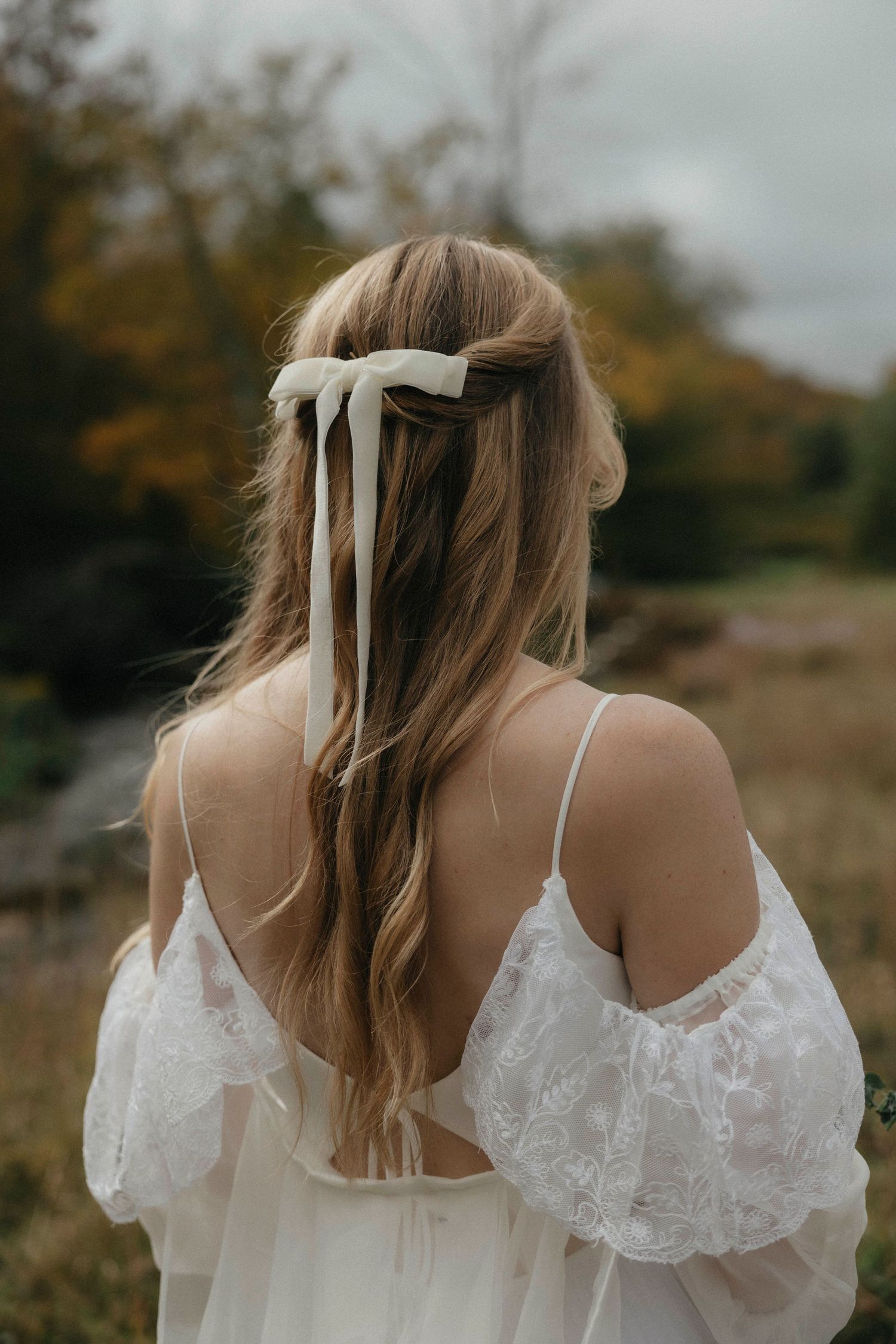 Back view of bride with long wavy hair adorned with white ribbon bow in lace dress.