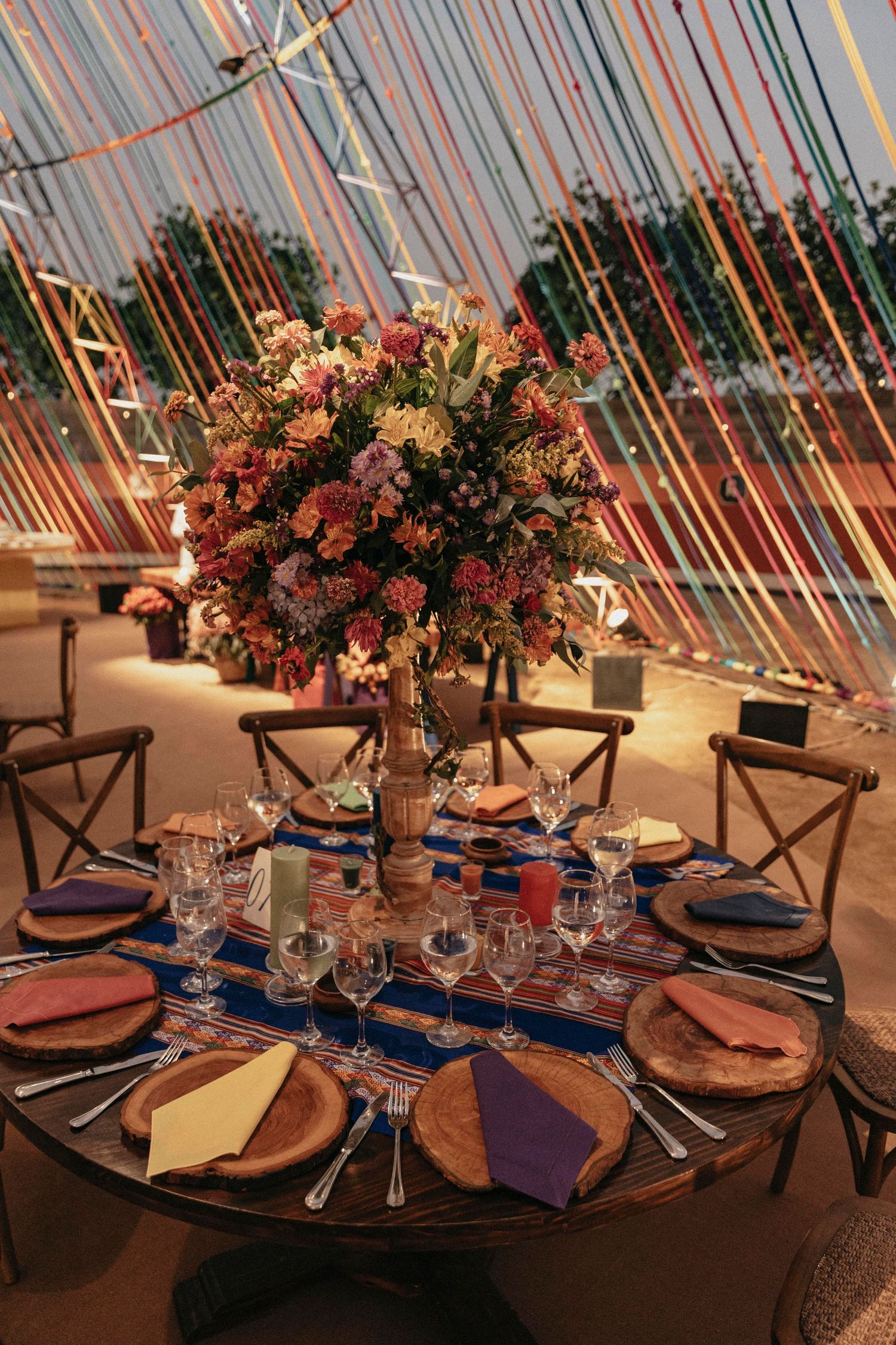 Elegant wedding reception table with tall floral centerpiece, colorful napkins, and rustic charger plates under string lights.
