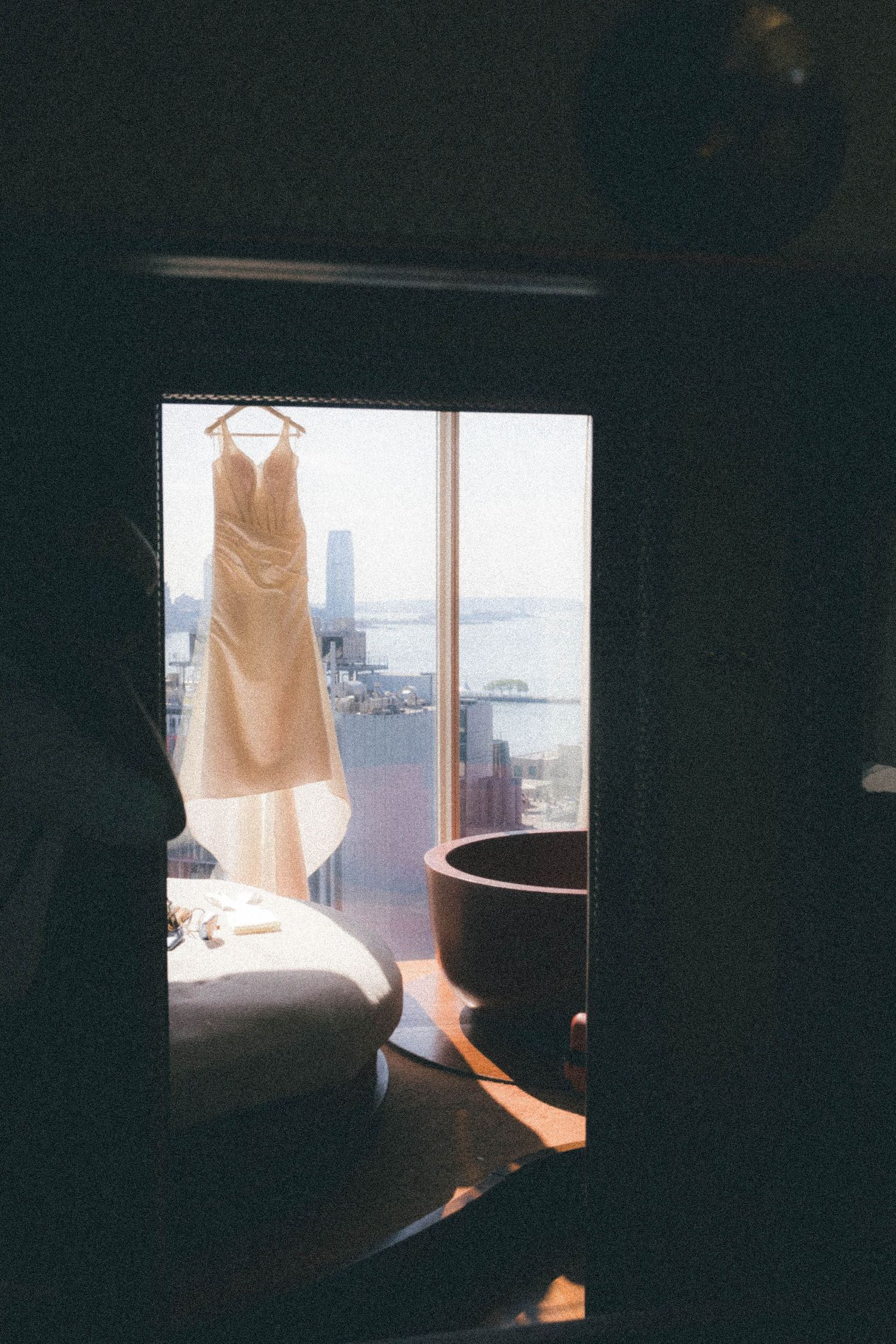 Wedding dress hanging on hanger in sunlit window with city waterfront view beyond.