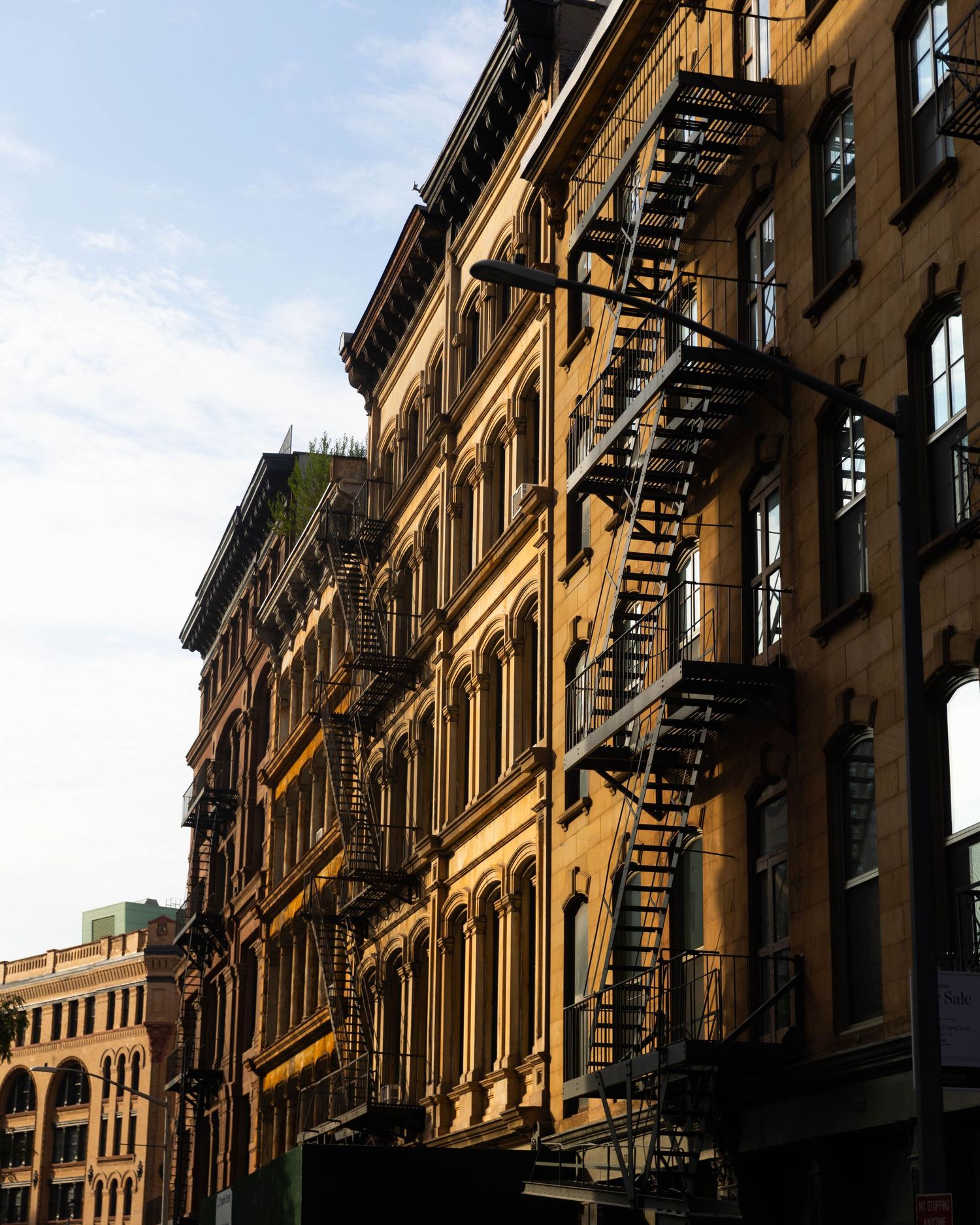 Sunlit brick buildings with tall arched windows and black iron fire escapes climbing the facades against a blue sky.