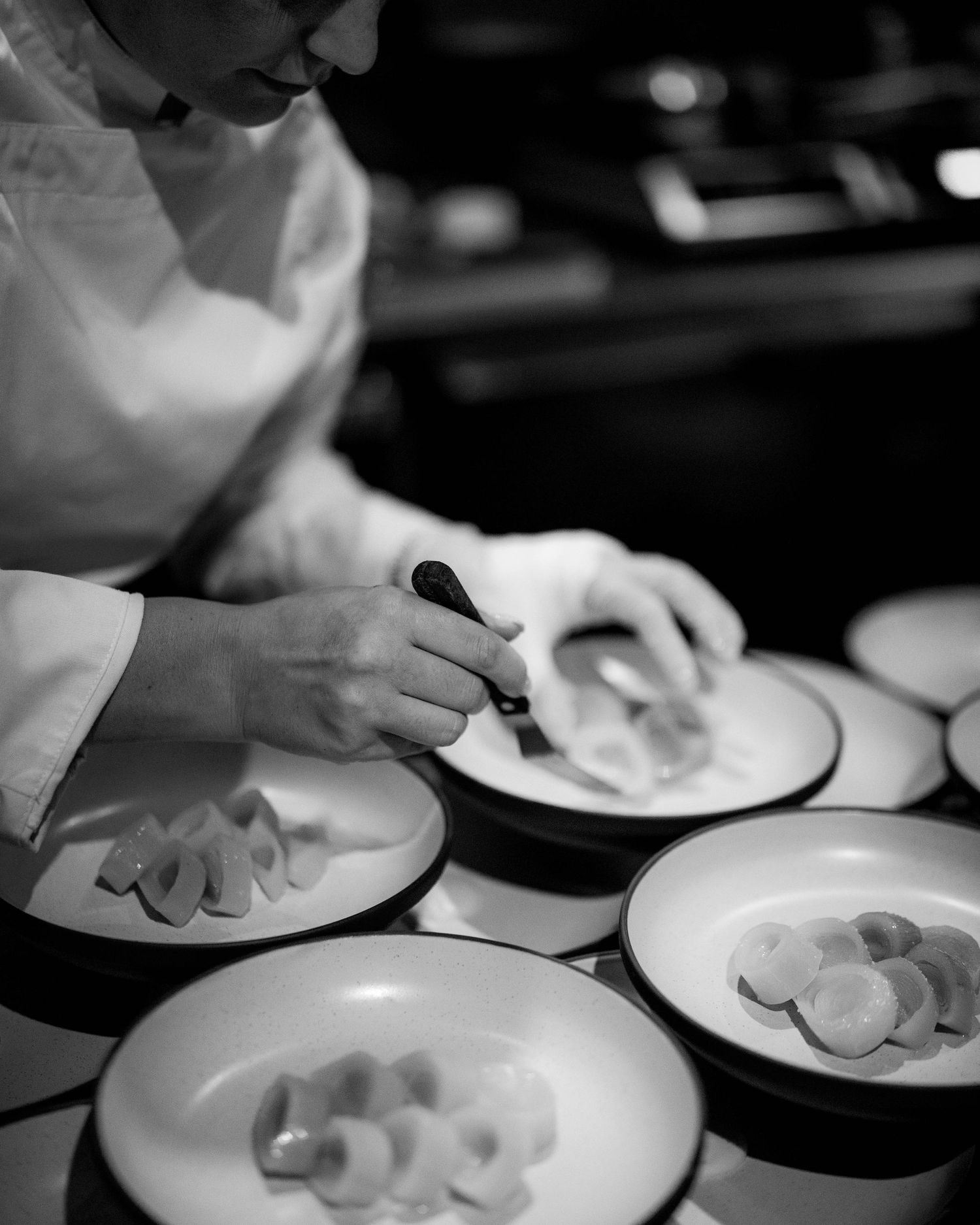 Black and white photo of a chef in whites using tweezers to carefully plate delicate sliced fruit on rows of rimmed ceramic plates.