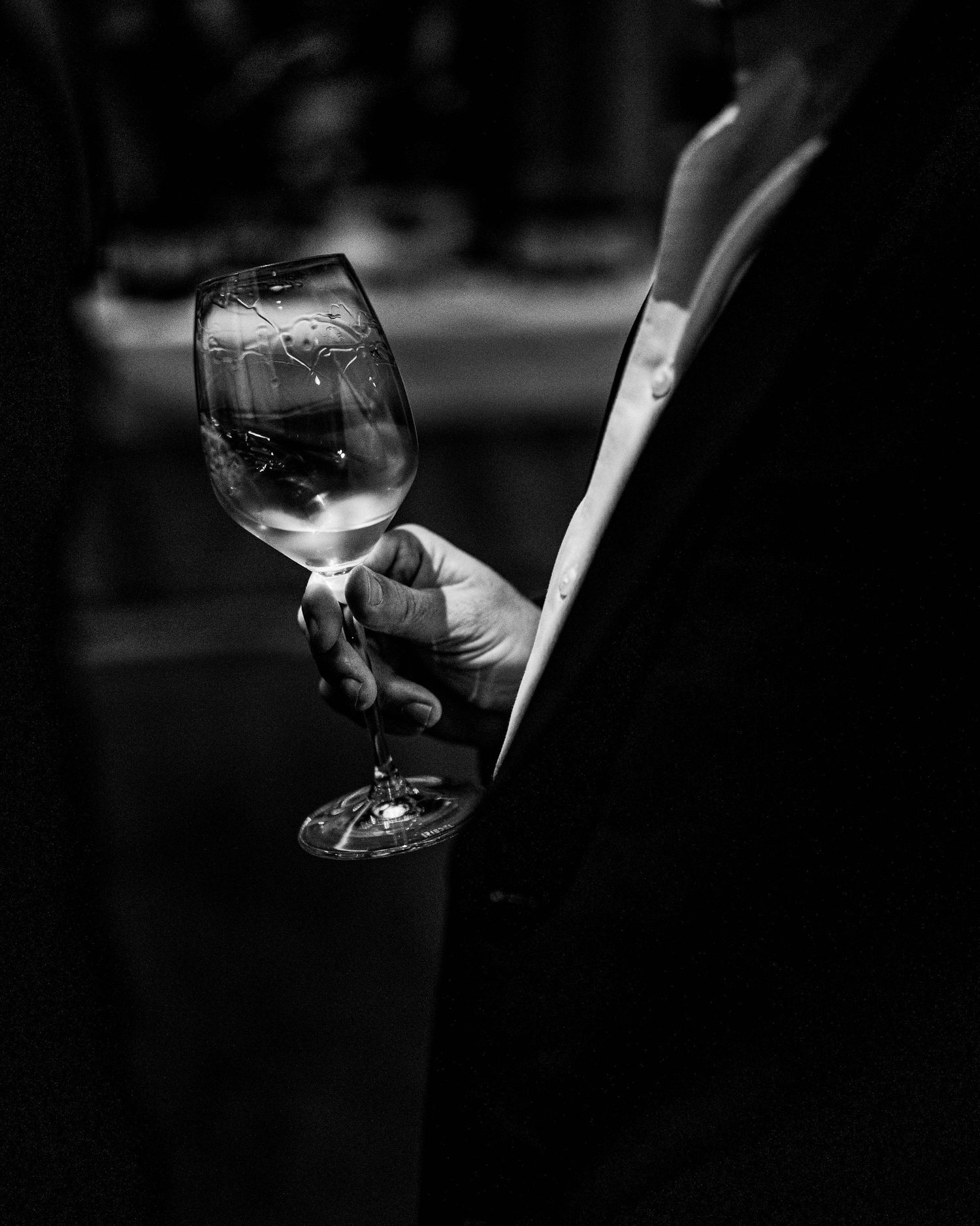 Black and white photo of a person in a dark suit holding a half-full wine glass during a wine tasting.