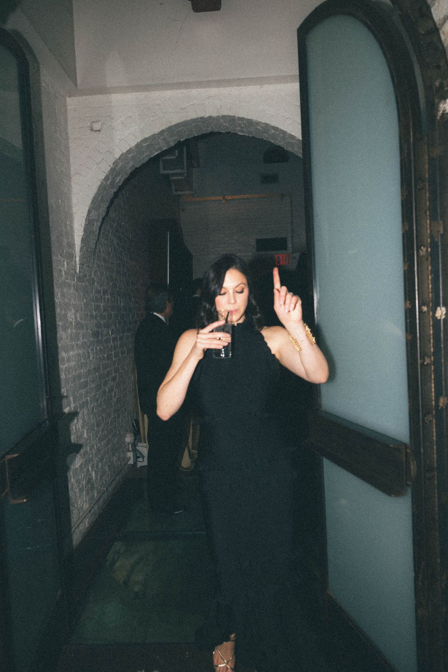 A woman in a black dress holds a cocktail while gesturing with her finger in an arched hallway.