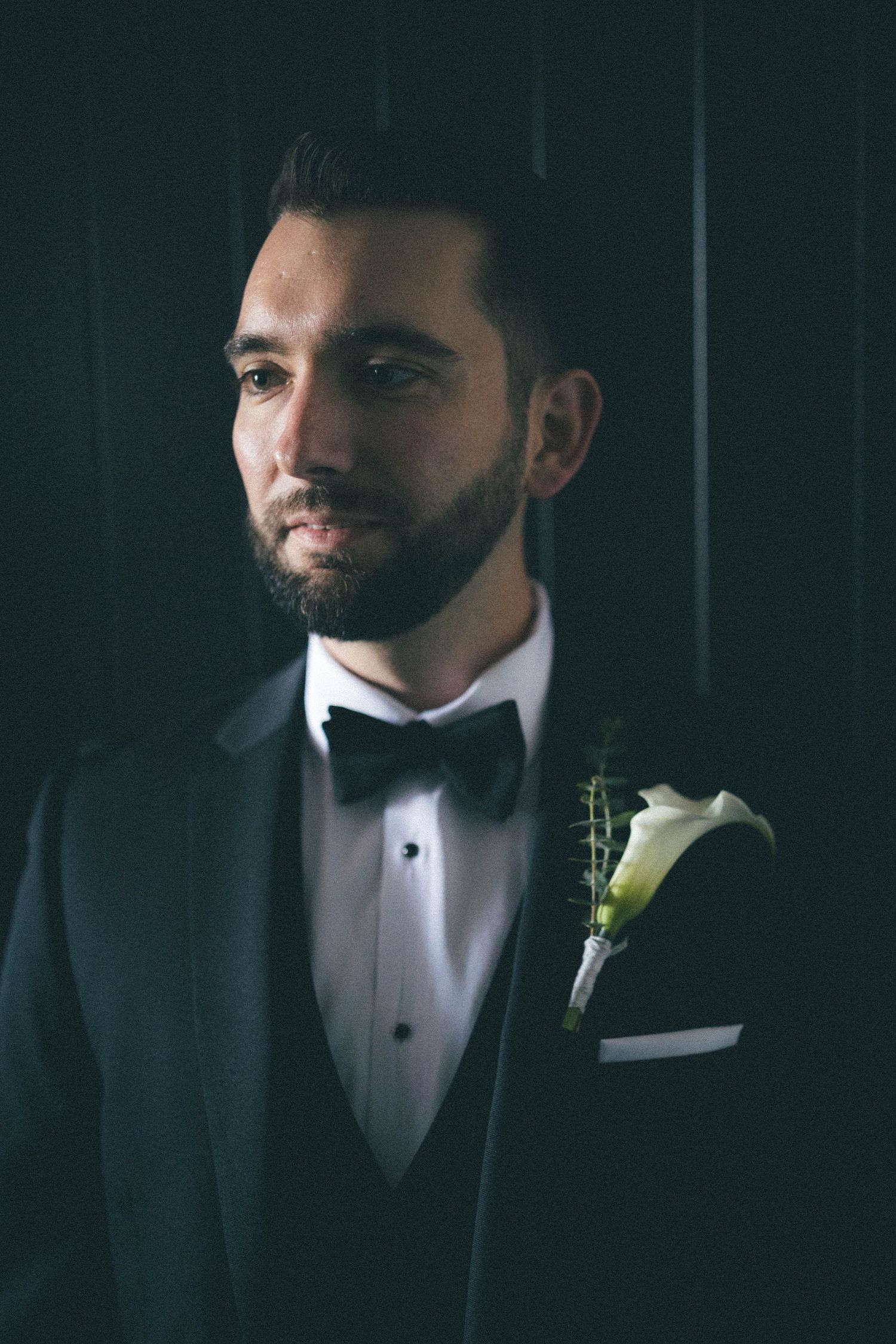 Handsome groom in black tuxedo with bow tie and white calla lily boutonniere against dark background.