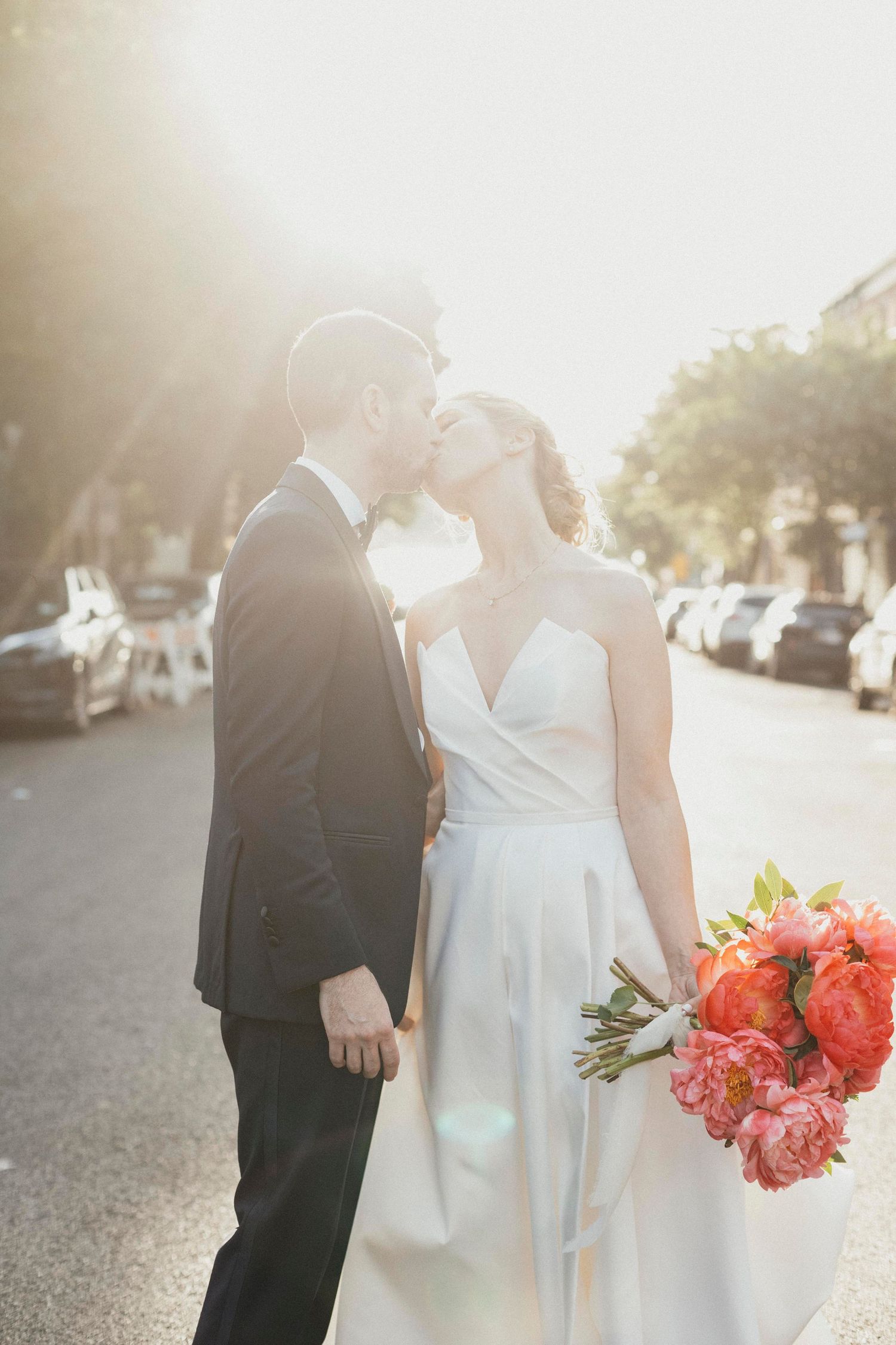 Bride and groom sharing a sun-kissed street kiss, bride holding bright coral peony bouquet in golden light.
