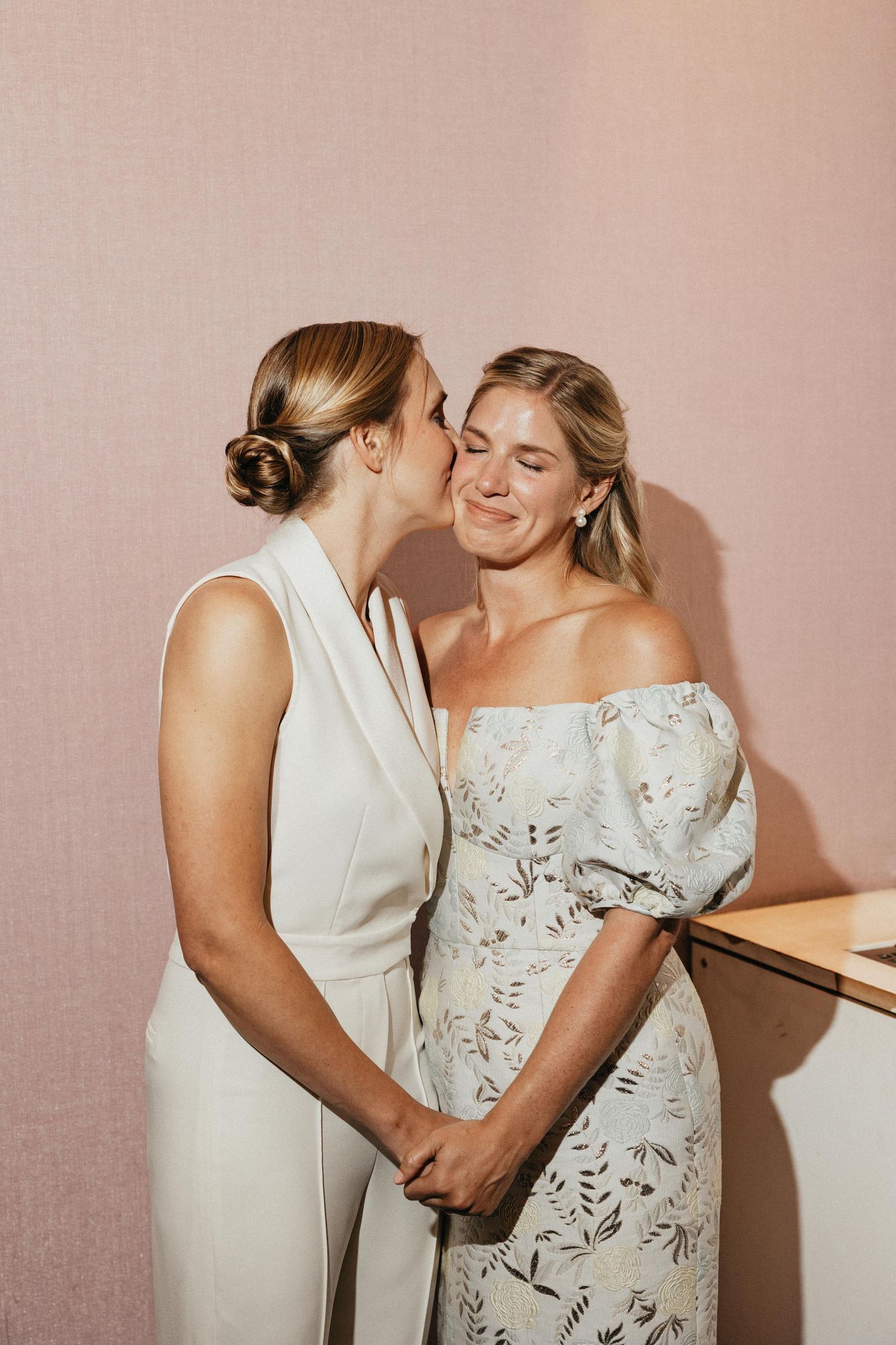 Two brides sharing a tender kiss on the cheek while holding hands against a pink wall.