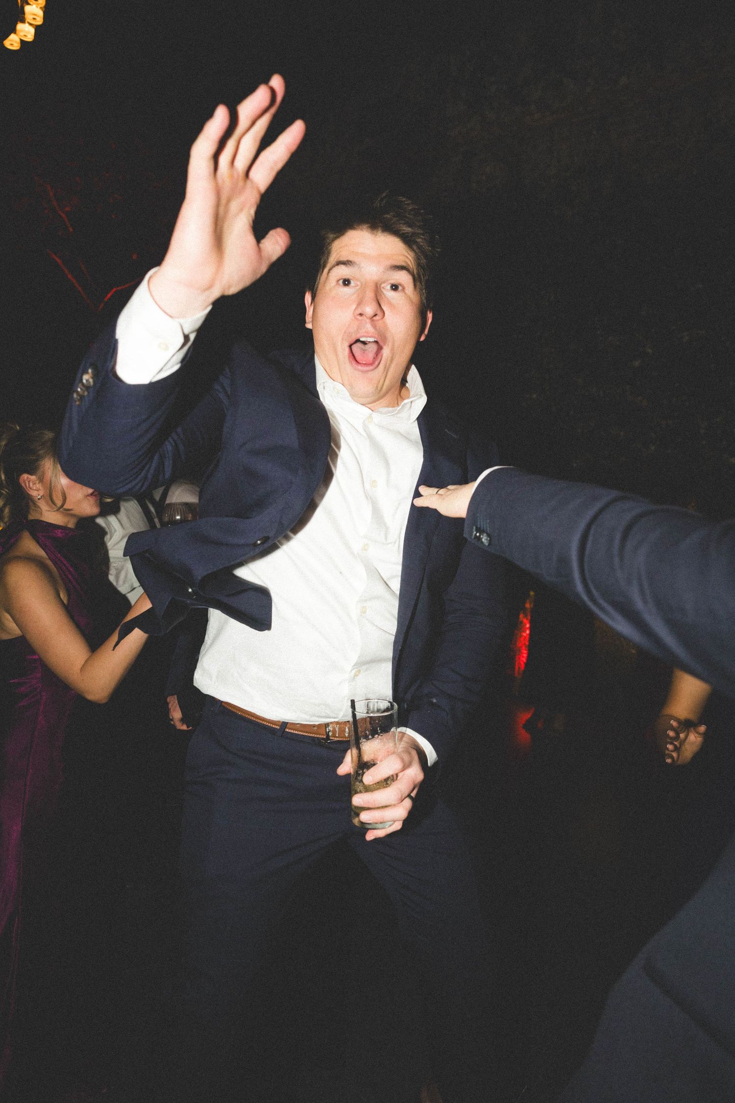 Energetic wedding guest in navy suit dancing with drink at lively wedding reception.