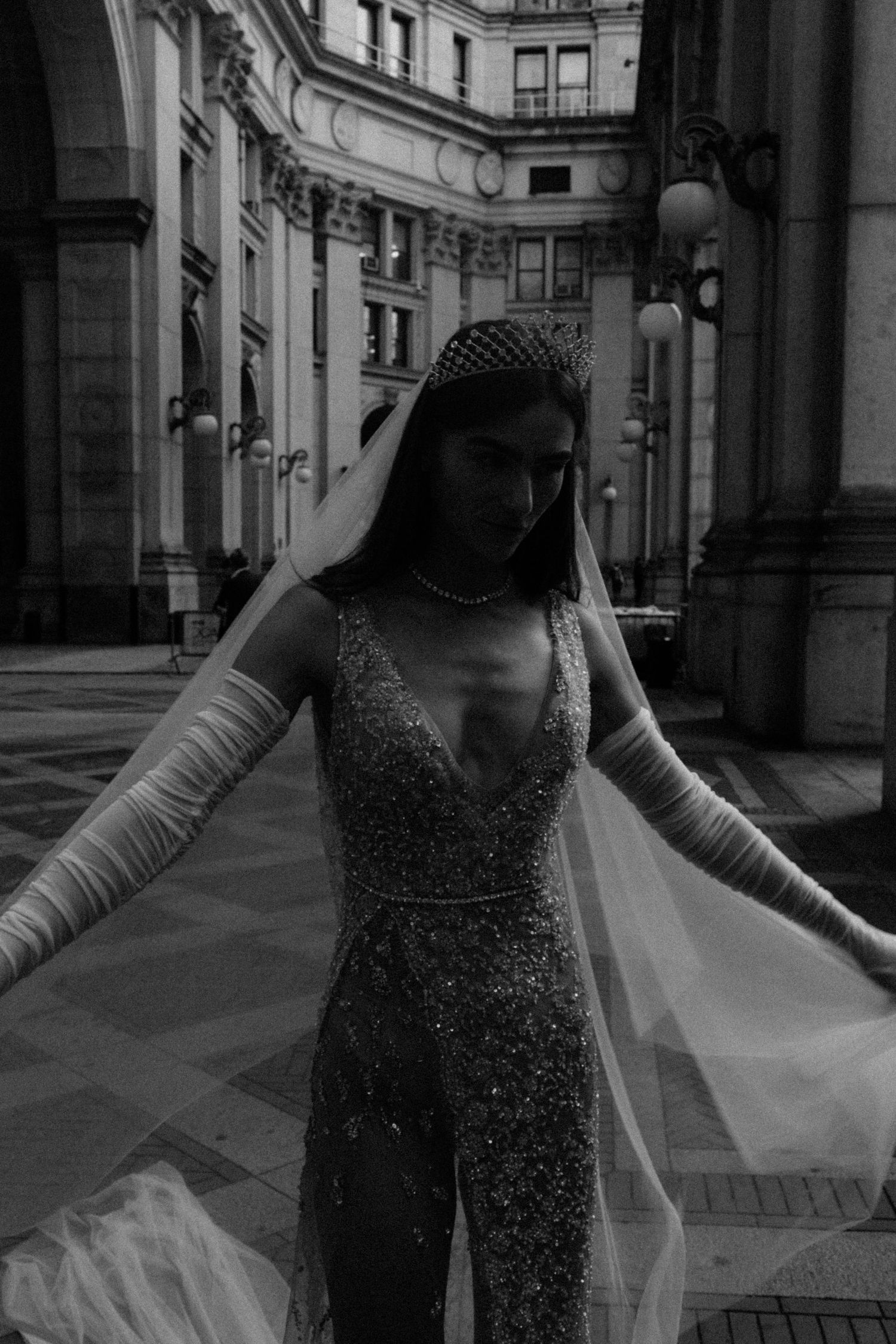 Black and white artistic photo of bride in beaded gown with mesh veil posing dramatically in a grand courtyard.
