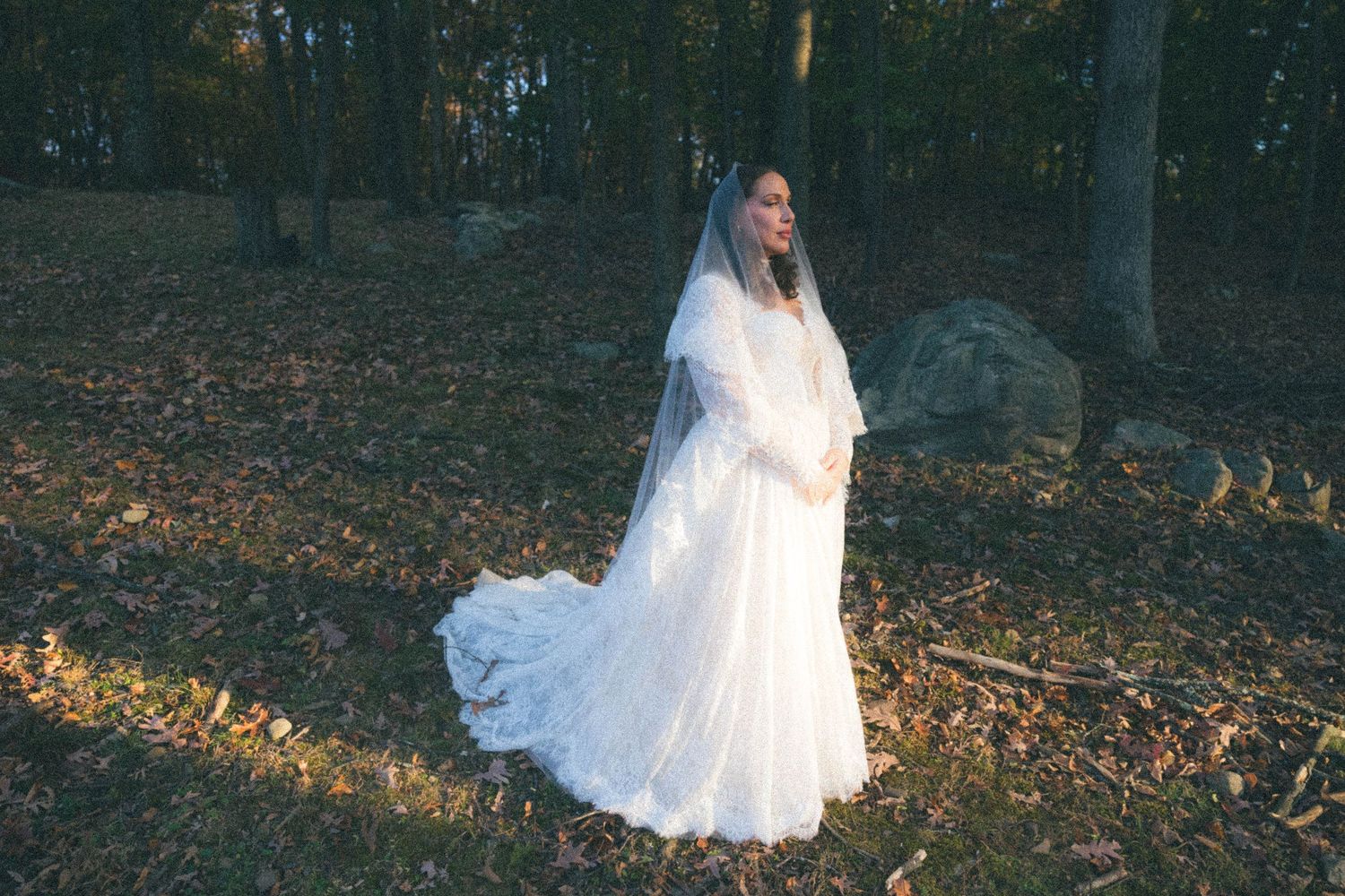Bride in white lace wedding gown with cathedral veil standing in autumn woodland forest setting.