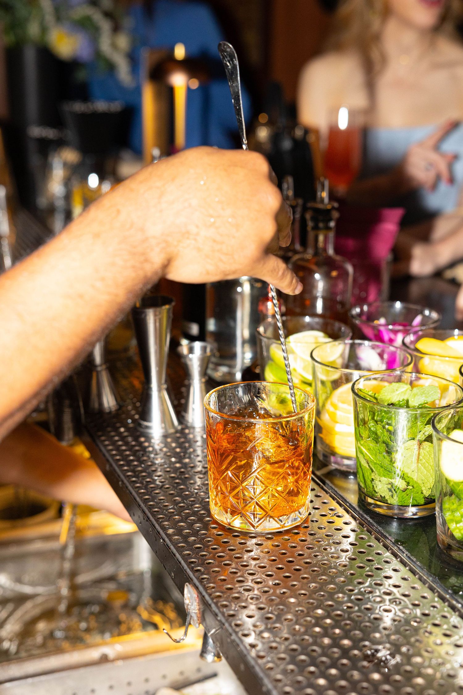 Bartender stirring amber cocktail on ice at bar with mojitos and other drinks in background.