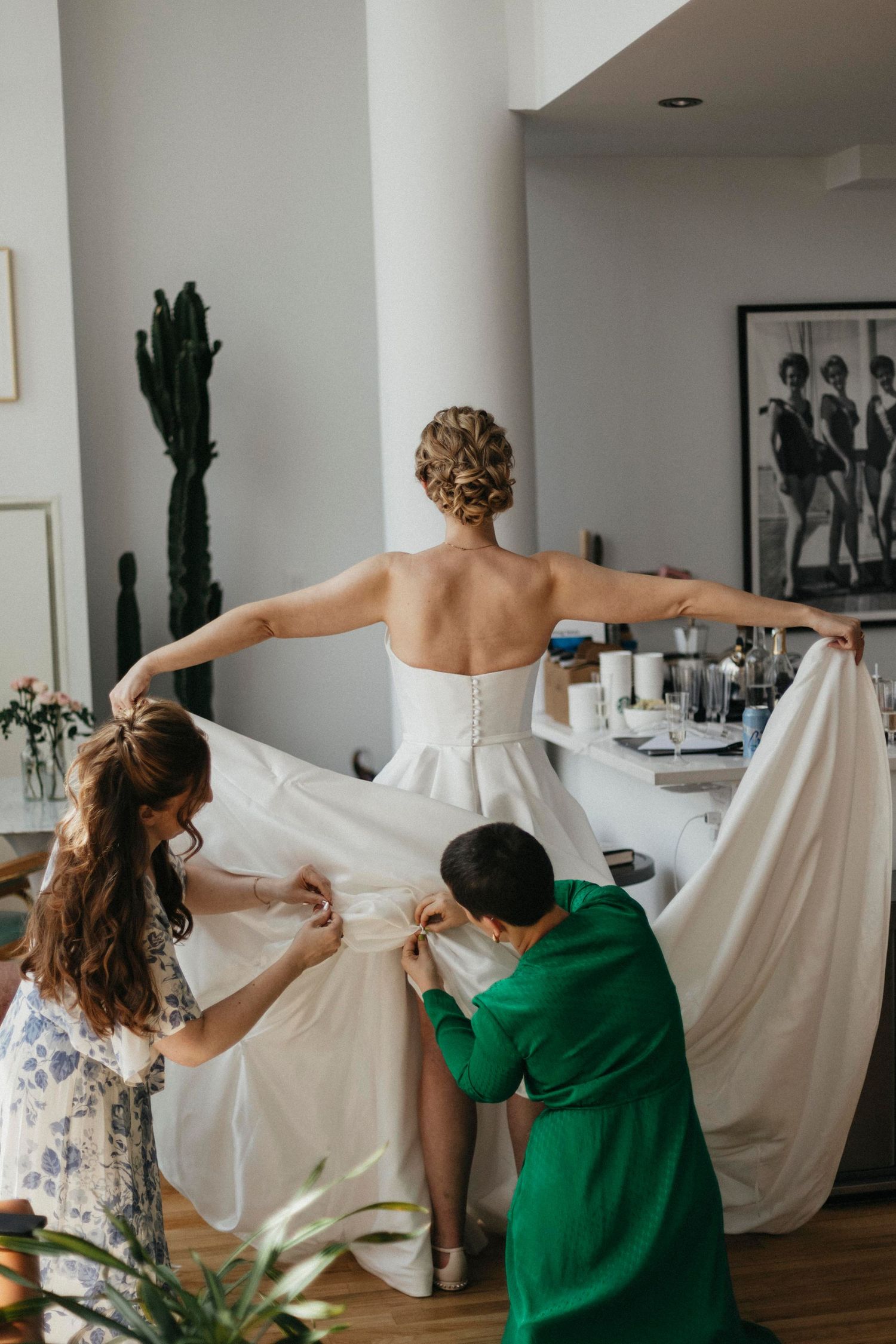 Bride with arms outstretched as two bridesmaids in floral and green dresses arrange her white wedding gown train.