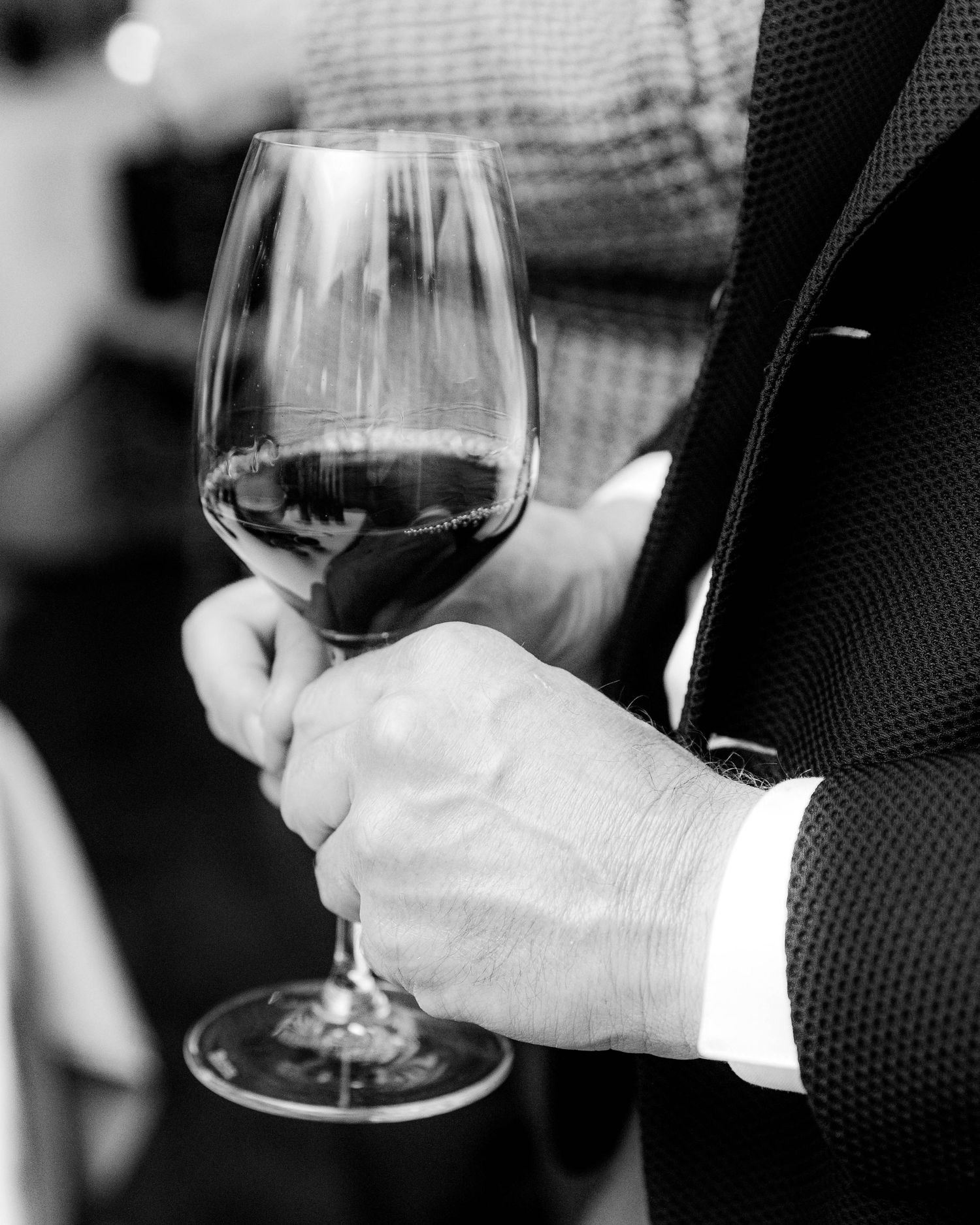 Black and white photo of a man in a textured suit holding a glass of red wine at an elegant event.