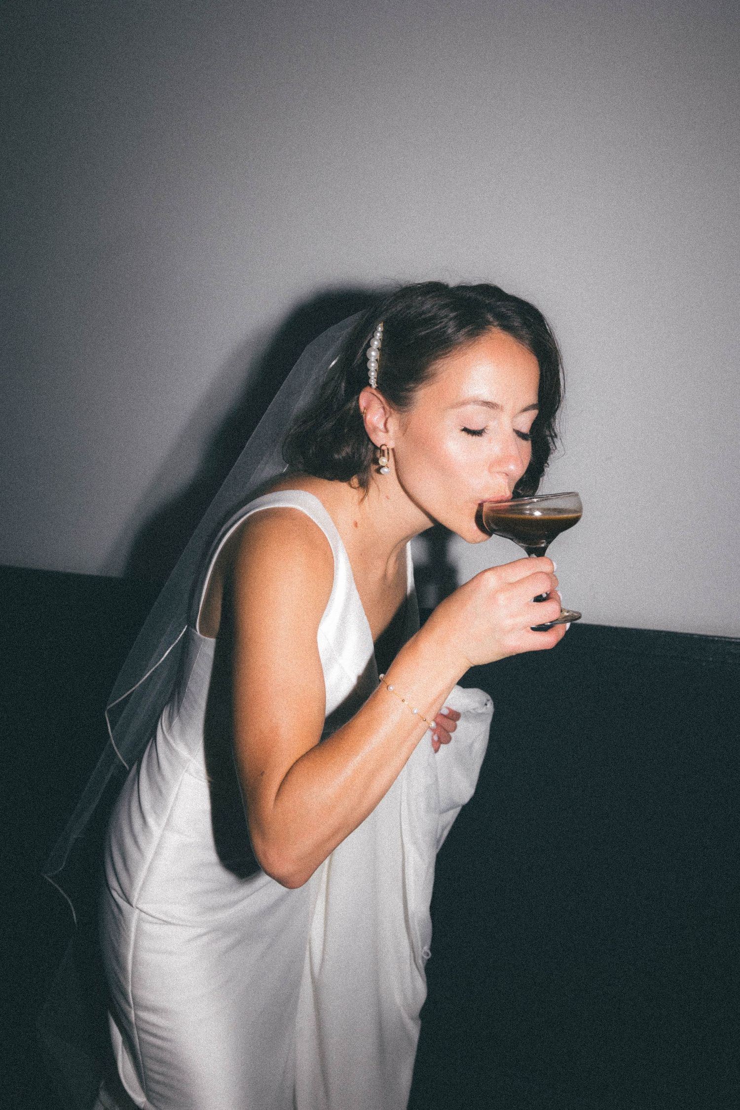 Bride in white gown and veil sips from a cocktail glass at her wedding reception.